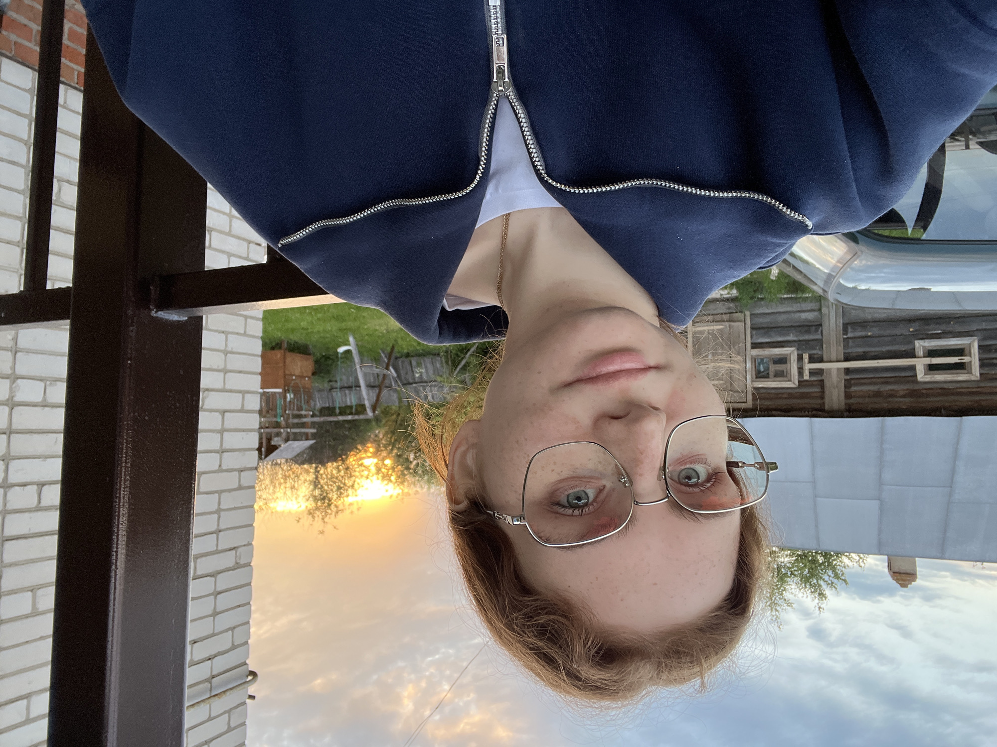 A young person, appearing to be female, takes a selfie outdoors, captured from an upward angle and presented upside down. She has light-colored hair, visible freckles, and wears silver-rimmed glasses. Her eyes are light-colored, possibly blue or green, and she has a neutral to slightly pensive expression. She is wearing a dark blue zip-up hoodie over a white t-shirt, with a thin gold chain necklace visible around her neck.
The background, when viewed correctly (rotated 180 degrees), shows an outdoor setting during what appears to be a sunrise or sunset, indicated by the warm, golden light on the horizon on the left side of the frame. The sky is partially cloudy, with soft light. To her left, there's a dark brown metal structure, possibly a railing or support beam, next to a white brick wall. Further in the background, there's green foliage, suggesting trees or bushes. On her right, a portion of a vehicle, possibly a car, is visible, reflecting the sky. Beyond that, a rustic wooden building with small windows can be seen. The overall setting suggests a residential or semi-rural area. There is no visible text in the image.