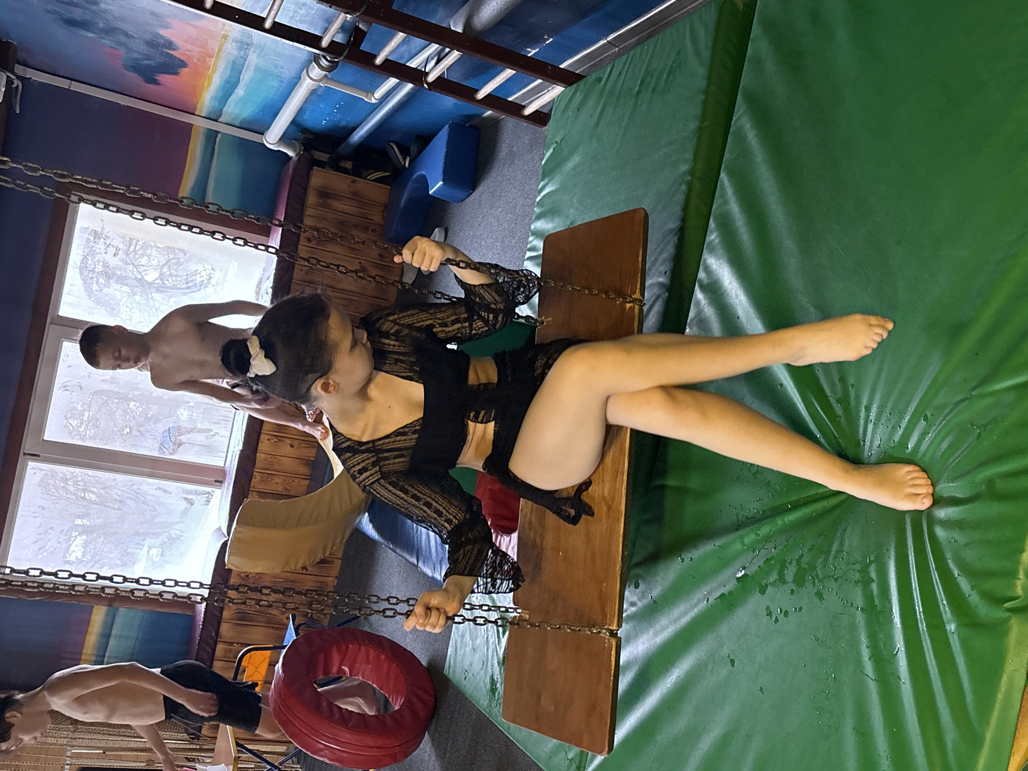 A medium shot depicts a juvenile female wearing black lace clothing sitting on a wooden plank swing suspended by chains inside a room. The female's legs are outstretched over a green padded mat. Her bare feet rest upon the mat. The female holds onto the chains with her hands. A juvenile male stands behind her, shirtless. Another juvenile male is partially visible in the lower left corner, shirtless. The background includes a window with a snowy view, a red tire-like object, and a padded blue cushion. The ceiling has painted decorations. The location is Unknown, Unknown.