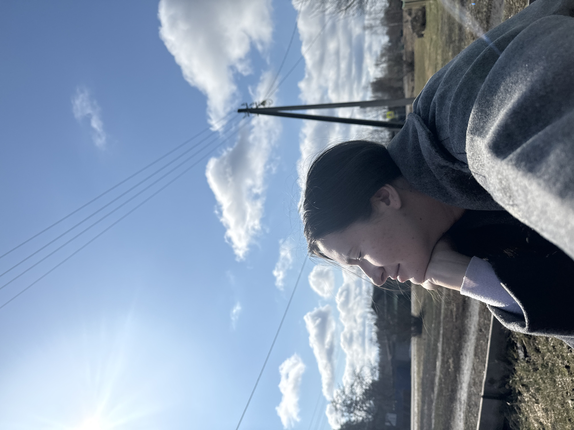 A woman is resting her chin on her hand, looking downward. She is wearing a gray coat with a lavender sleeve visible underneath a darker jacket. The sky is blue with white clouds and a bright sun in the lower left corner. Power lines and a utility pole are visible in the background. A fence or wall runs along the bottom right of the image, with vegetation and trees beyond. The location is outdoors.