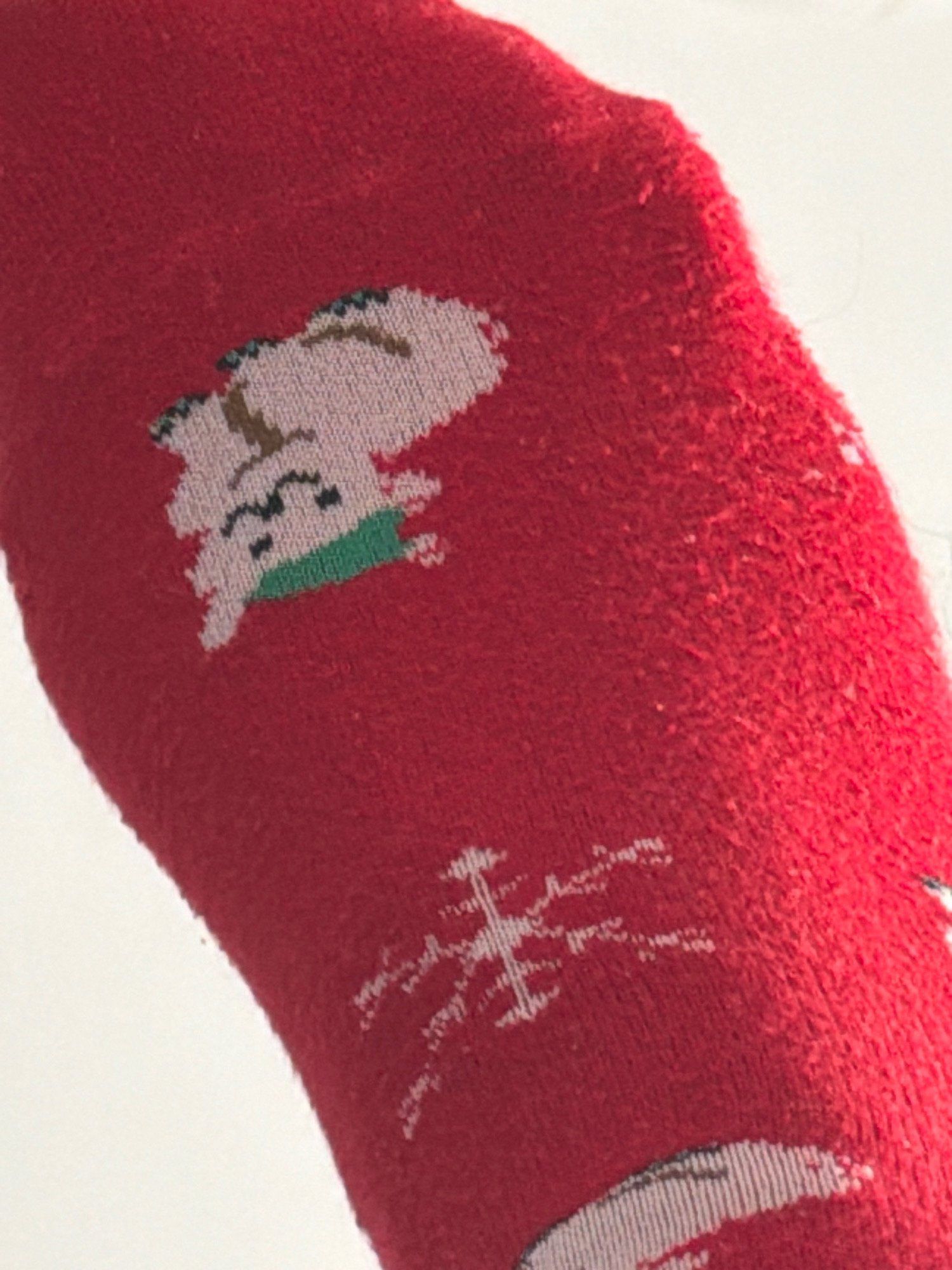 The image shows a close-up of a red knitted sock. The sock has a pattern of cartoon characters, possibly Christmas-themed, knitted into the fabric. One character appears to be a light-colored animal with brown accents and a green scarf. Another element of the pattern is a white, snowflake-like design. The fabric of the sock appears slightly pilled. The background is a plain white surface. There is no indication of the sock's location in Khabarovsk, Russia, or any other geographical context.