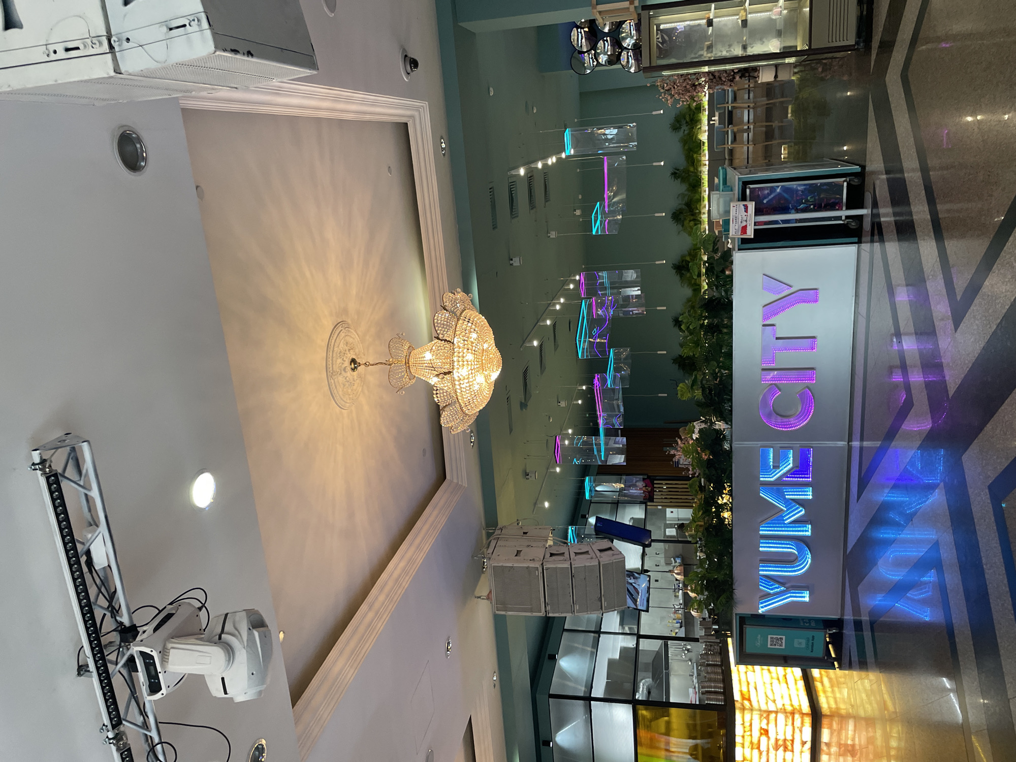 This is a low-angle interior shot of a modern establishment, likely a restaurant or entertainment venue, with a focus on the ceiling and a prominent sign. The ceiling features intricate crown molding, a large ornate chandelier, and various lighting fixtures, including a bar light and what appear to be stage lights suspended on a truss. Multiple speakers are mounted overhead, suggesting a sound system for events or ambient music. Hanging from the ceiling are several translucent, rectangular light fixtures emitting a vibrant blue and purple glow. The main subject of the image is a large, illuminated sign spelling out "YUME CITY" in large, block letters that transition from bright blue on the left to purple on the right. The sign is mounted on a white counter that runs horizontally across the foreground. The floor is a polished, reflective dark surface with geometric patterns. Behind the counter, glimpses of a kitchen area with stainless steel appliances, dining tables with chairs, and lush green plants are visible, hinting at the venue's purpose. The overall ambiance is modern and vibrant, with a strong emphasis on lighting design.