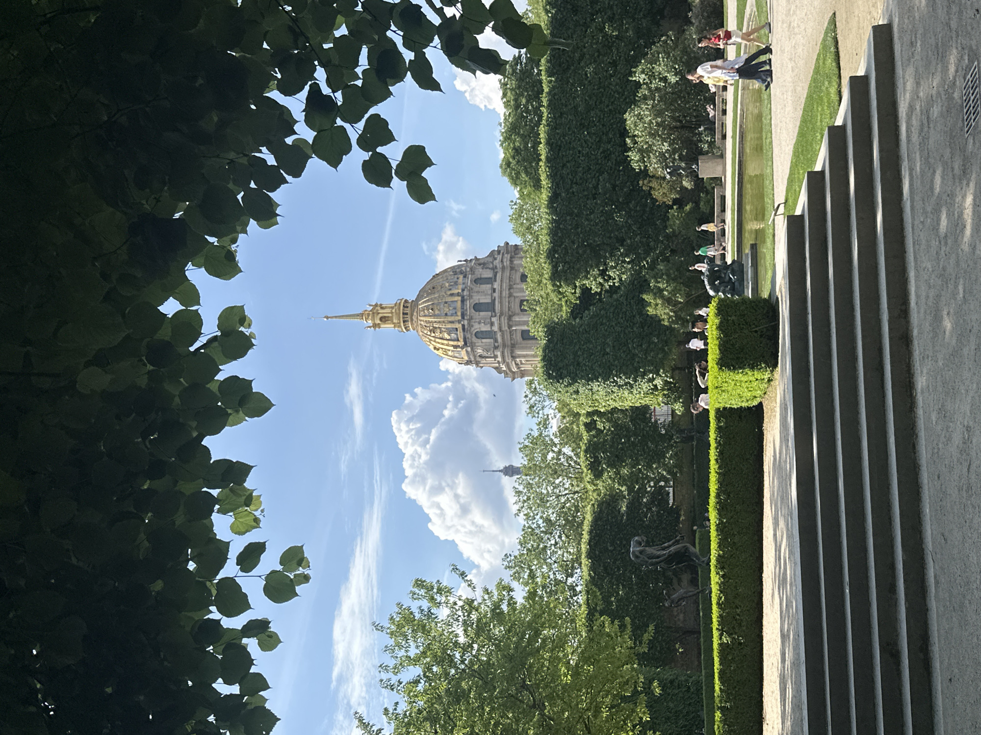 The image captures a view in Paris, France, framed by trees. The dome of the Invalides is prominently visible, rising above manicured hedges and greenery. The Eiffel Tower is also visible in the background, smaller and further away. On the right side of the frame, concrete stairs are visible, and people can be seen walking near a reflecting pool and a garden. The sky is blue with scattered clouds.