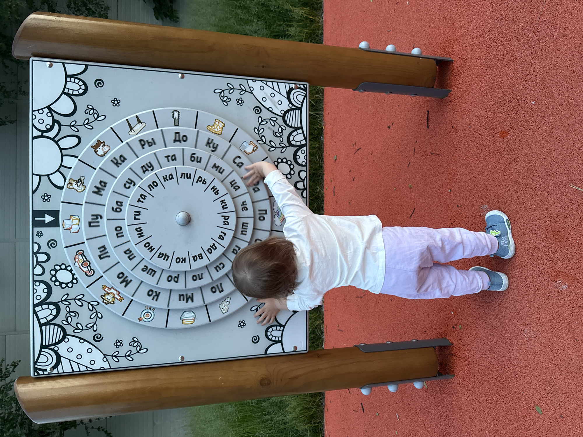 In Moscow, Russia, a young child, viewed from behind, stands on a red rubberized surface in a playground. The child, wearing a white long-sleeved shirt and light purple pants, has their hands outstretched towards a large, circular interactive game panel. This panel, encased in a wooden frame, features concentric rings with images and Cyrillic letters, designed for children's educational play. The background shows a blurry green area, suggesting natural vegetation.
