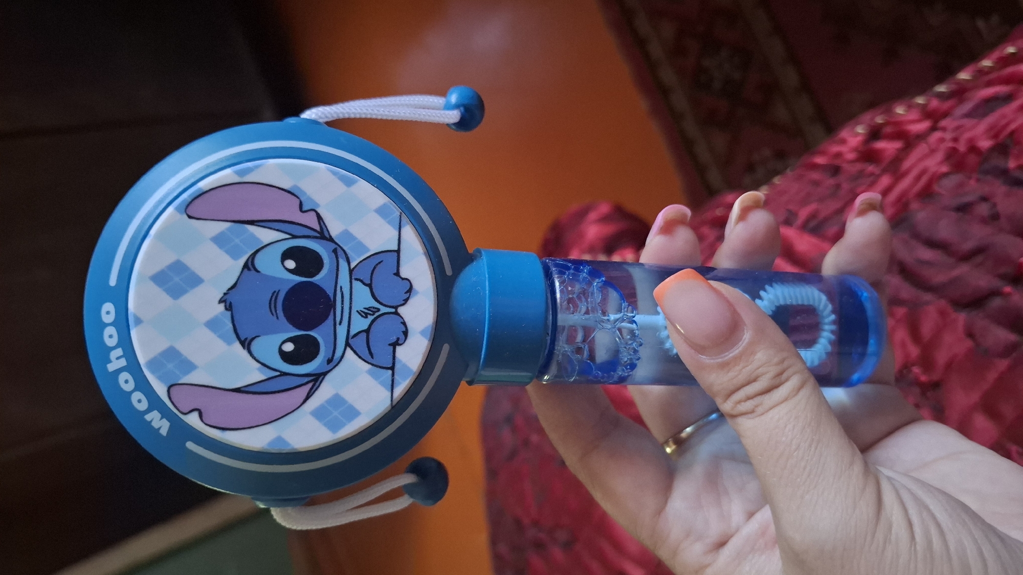 A person's hand is holding a blue bubble wand with the character Stitch from Disney's Lilo & Stitch on it. The wand has a round blue head with a picture of Stitch and the word "Woohoo" written in white. There are white strings with blue beads attached to the sides of the head. A small blue bottle filled with bubble solution is attached to the bottom of the wand. The background is blurry and appears to be indoors with a reddish-brown surface. The image is taken from a close-up perspective, focusing on the bubble wand and the hand.