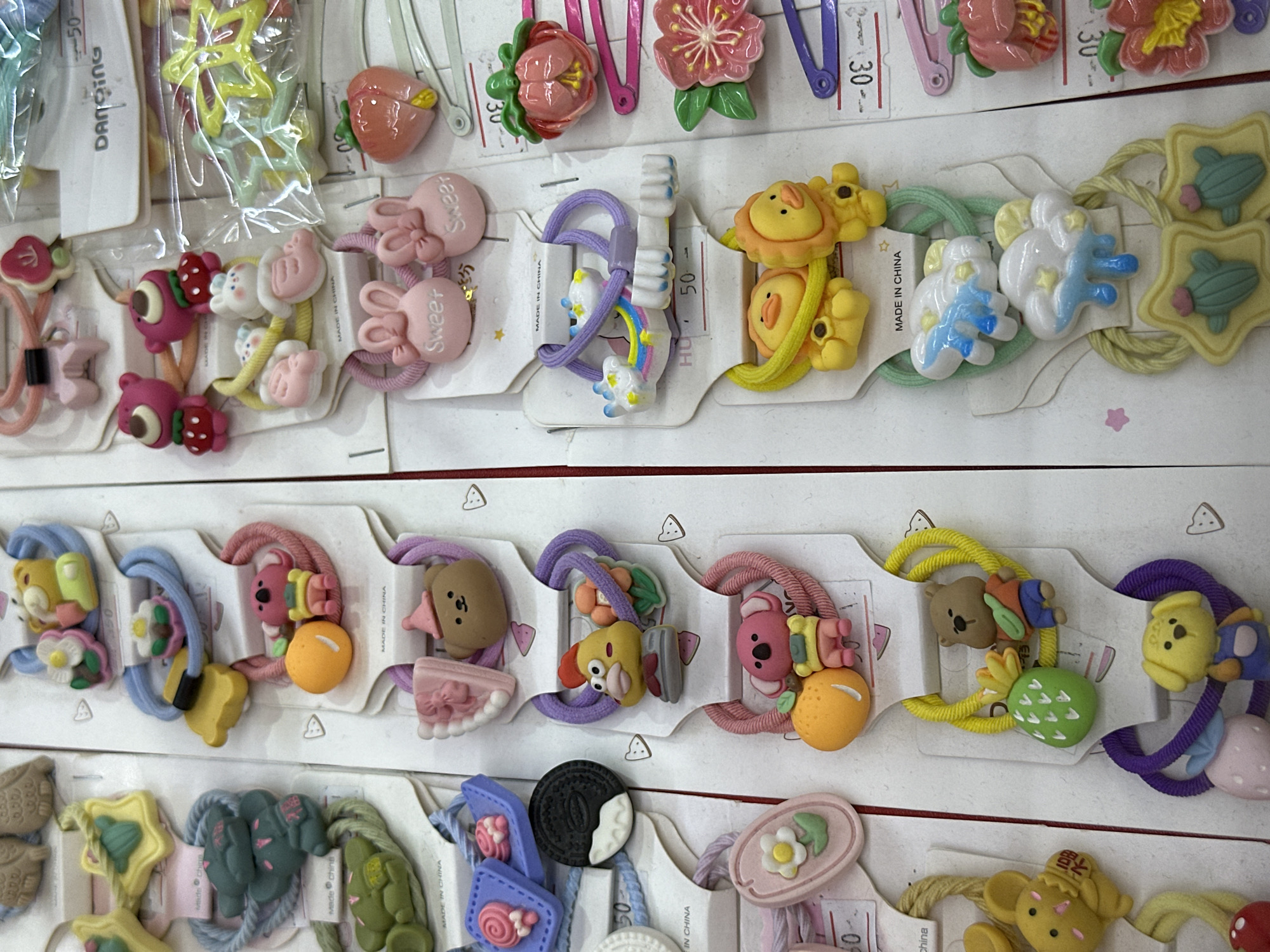 This media file is a close-up, overhead shot of various hair accessories, primarily hair ties and clips, arranged in rows. The items are colorful and predominantly feature cartoon-like animal figures, fruits, and flowers.
The subjects are objects, specifically hair accessories. They include pink bunny-shaped hair ties, hair ties with strawberry and other fruit charms, bear-shaped hair ties, and hair clips adorned with colorful flower designs. Some hair ties have small, detailed charms like a smiling lion, a yellow bird, and clouds with rainbows. There are also hair ties with orecchiette pasta-shaped charms and a single oreo cookie-shaped charm.
The setting appears to be a retail display or a close-up view of a collection of these items, likely for sale. The items are presented on white cardstock backing, which is characteristic of retail packaging for such accessories. The background is a neutral, white surface, suggesting a clean, organized display.
There are no people visible in the media file. The activity, if any, is simply the presentation of these merchandise items.
Notable details include the visible text on some of the packaging, such as "MADE IN CHINA" and "DANCING". Some Chinese characters are also visible on one of the yellow hair ties, which translates to "fortune" or "blessing". The price "50" is also visible on some tags. The overall impression is one of a vibrant and playful collection of children's hair accessories. The lighting is even and bright, indicating it could be under artificial light or in a well-lit environment. No time of day or weather cues are available. The location is inferred to be a retail setting, but the specific geographic location is unknown.