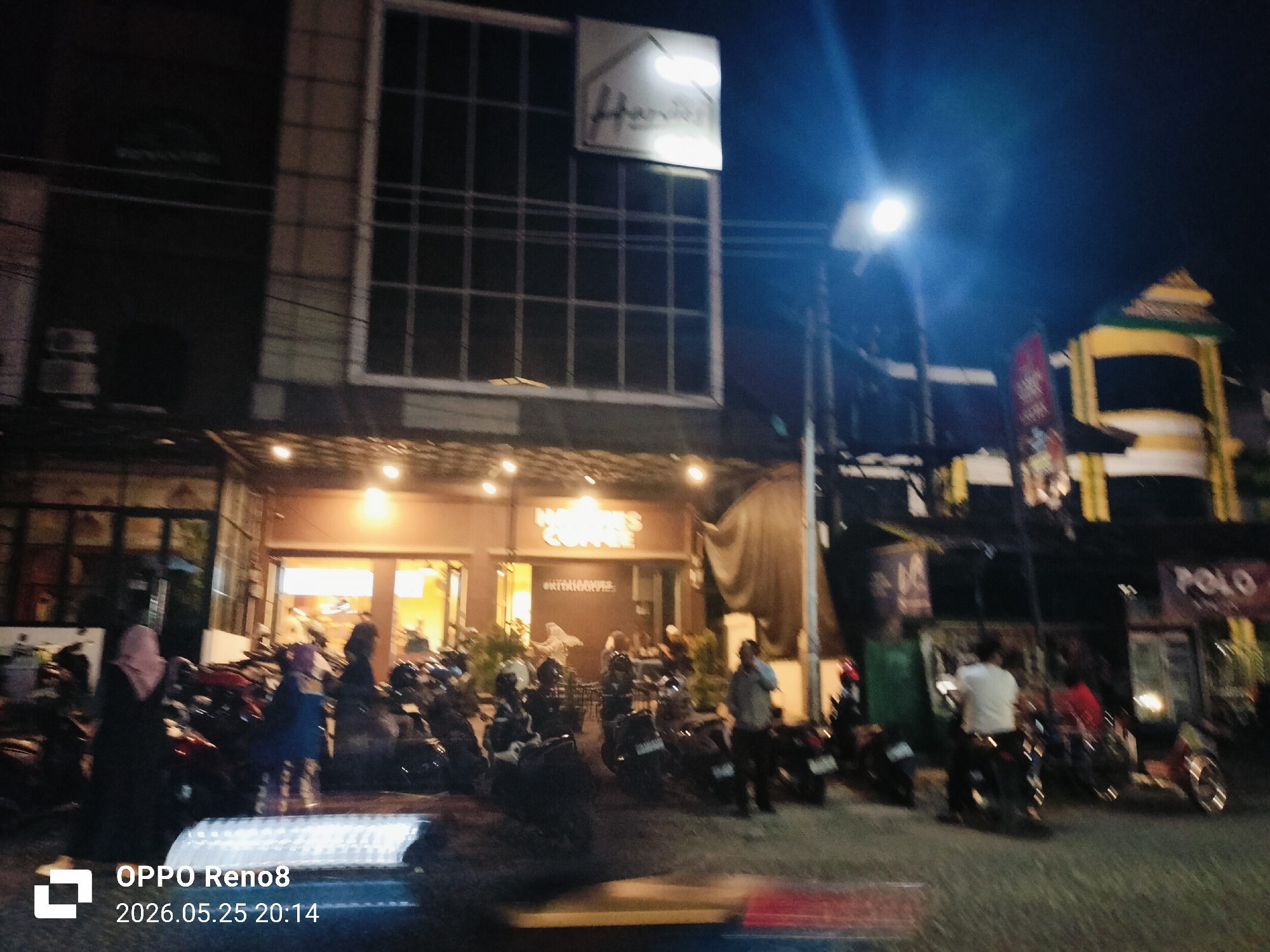 The image shows a street scene at night in Lhokseumawe, Indonesia. A two-story building with a sign that reads "Hanna's Coffee" is visible, with people seated at tables outside. Many motorcycles are parked in front of the establishment. The street appears busy, with pedestrians and people on motorcycles. A bright street light illuminates the scene. In the background, another building is visible with a sign that reads "POLO". The camera date stamp indicates the picture was taken on 2026.05.25 at 20:14.