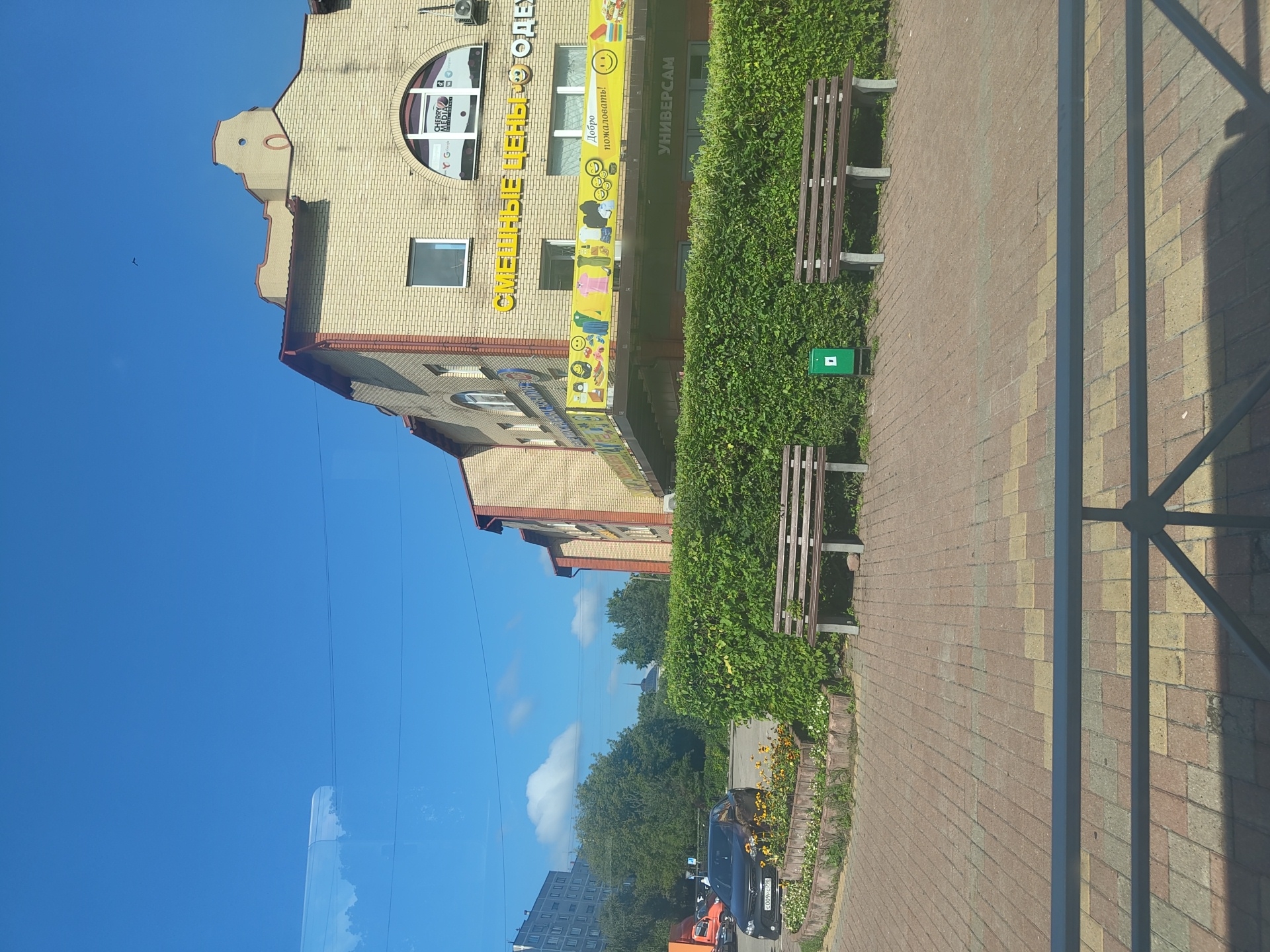 The image captures a sunny day in Ramenskoye, Russia, with a bright blue sky and scattered clouds. In the foreground, a paved walkway made of interlocking bricks leads towards a row of benches. Two wooden benches with concrete supports are positioned in front of a dense, green hedge. A green trash bin is located between the benches.
Behind the hedge, a light-colored, brick building stands tall. Yellow, raised lettering on the building facade reads "СМЕШНЫЕ ЦЕНЫ" (Funny Prices) and is accompanied by smiling emoji graphics. Below this, a banner displays cartoonish images of clothing items and more smiley faces, along with the Russian phrase "Добро пожаловать!" (Welcome!). Above this banner, a smaller sign for "CHERRY MEDIA" is visible. A large arched window with a semicircular top is also present on the building's facade.
To the left of the building, a dark-colored car with a license plate is parked. Further in the background, more trees and buildings are visible, including a tall apartment building and what appears to be a church spire. The overall scene depicts a commercial area with public seating, suggesting a retail or shopping district.