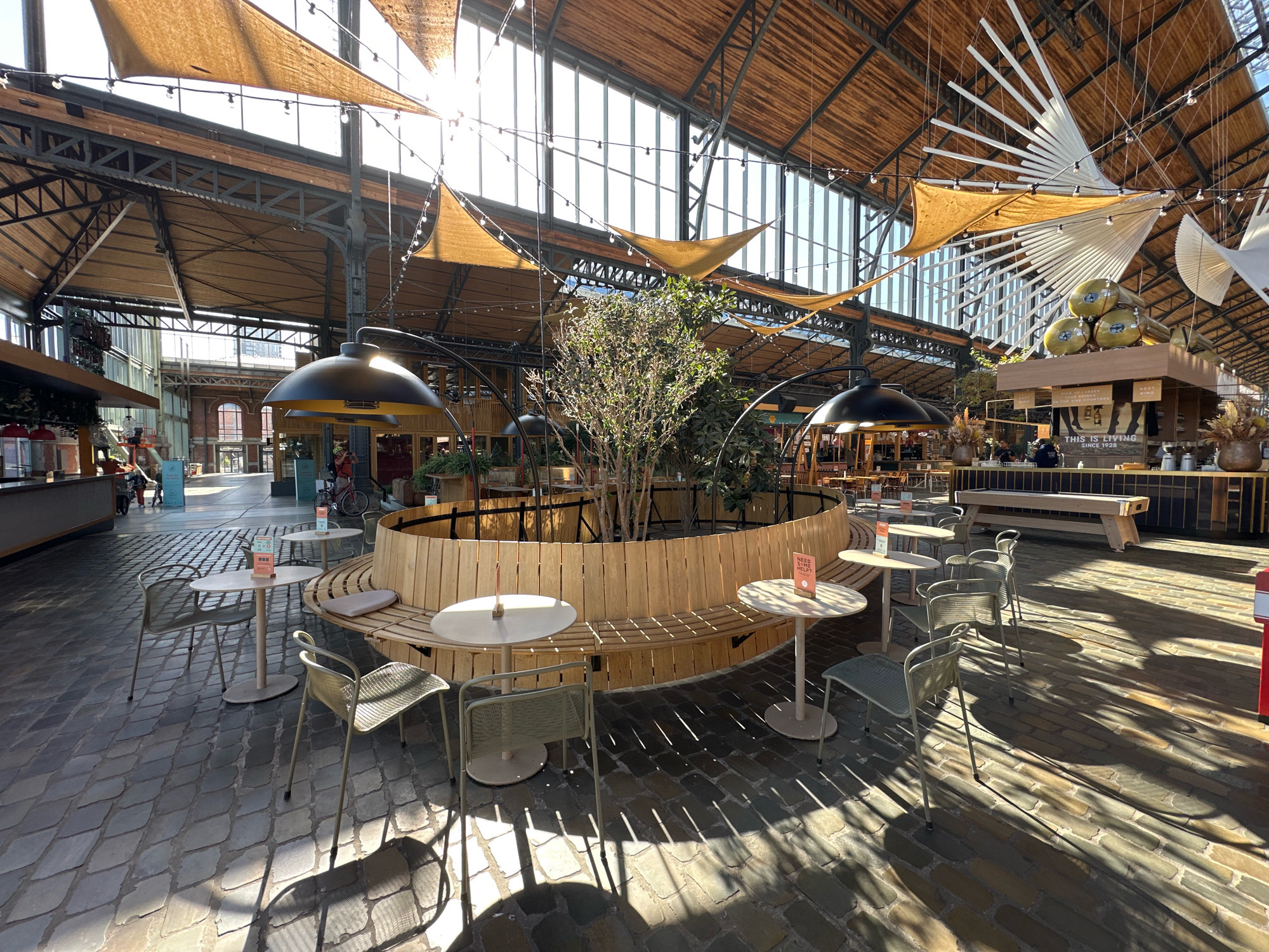 This image depicts the interior of the Brussels Central Station's new food court, located in Brussels, Belgium. The expansive space features a high, arched ceiling with exposed wooden beams and intricate metalwork. Large windows allow natural light to illuminate the area, supplemented by strings of decorative lights.
In the foreground, a circular seating area is defined by a light-colored wooden bench and several small, round tables. Two of these tables have chairs, one with a single chair and another with two. The tables are simple, light-colored discs on single pedestals. The chairs are modern, light green, metal chairs with a slightly curved design. The flooring is paved with dark, irregularly shaped stones, creating a textured surface.
The central element within the seating area is a large, potted tree or shrub with dense foliage. Surrounding this, the wooden bench curves around, providing ample seating. On one of the tables, a small pink sign with the text "NEED HELP?" is visible.
Further back, other seating arrangements and tables are visible. A long, wooden communal table with benches is situated in the middle ground. Behind this, a counter area is visible, with stools and a servery. Decorative elements, including large pendant lamps with black shades and abstract ceiling installations made of white and cream-colored materials, are prominent. Several large, golden-colored spheres are stacked on a display.
In the background, through large windows, a glimpse of the city outside is visible, including buildings and what appears to be a street. A few individuals are present, one person is standing near a counter, and another is walking past a bicycle in the distance. The overall impression is a modern, open, and inviting dining and social space within a historic building.