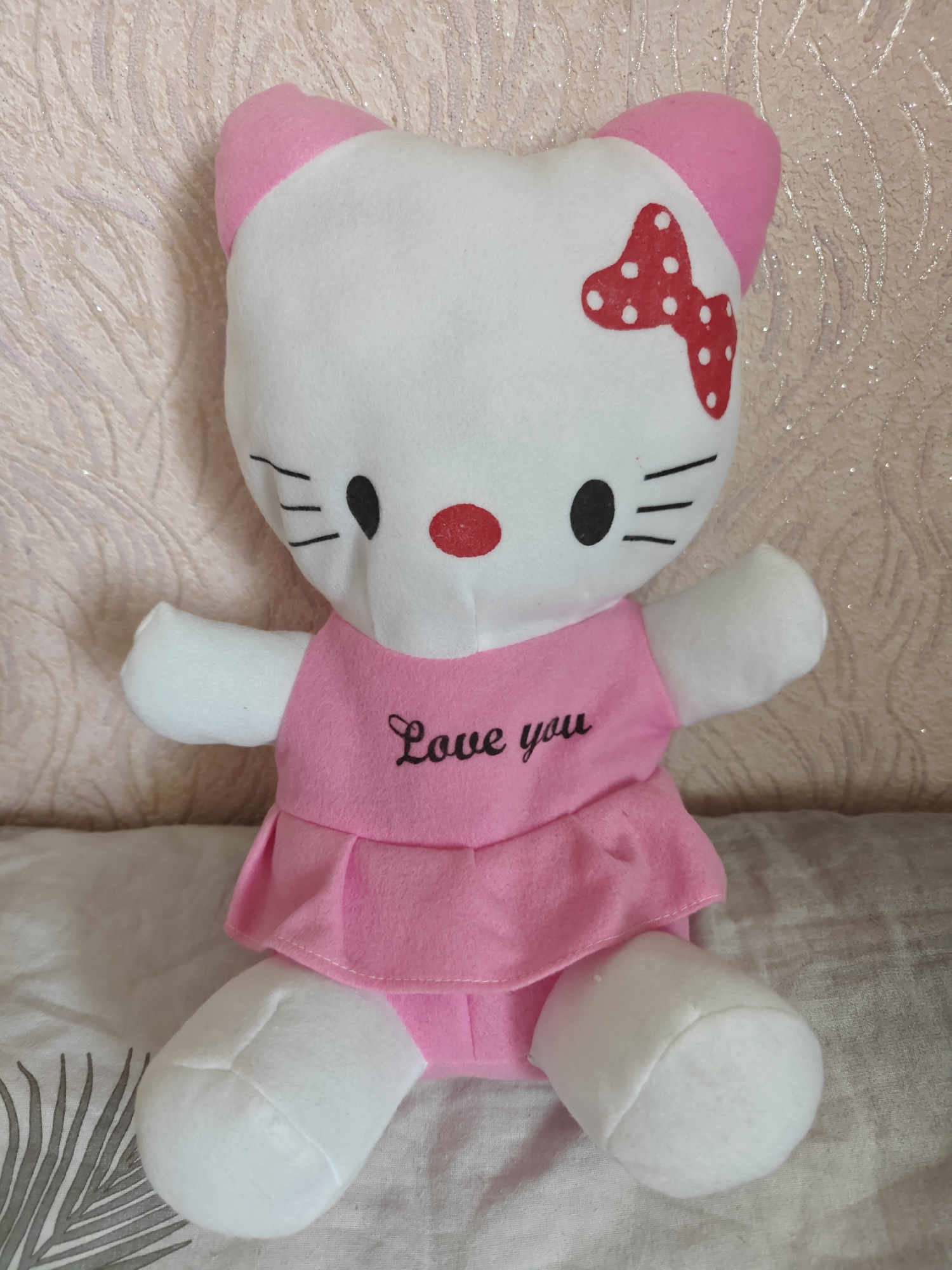 A plush toy resembling Hello Kitty is sitting on a bed. The toy is white with pink ears and a pink dress that says "Love you" in black cursive. It has a red bow with white polka dots on its head. The background is a textured, pale pink wallpaper. The bed has a white sheet with a gray palm leaf pattern. The image does not contain information about its location in Rubtsovsk, Russia.