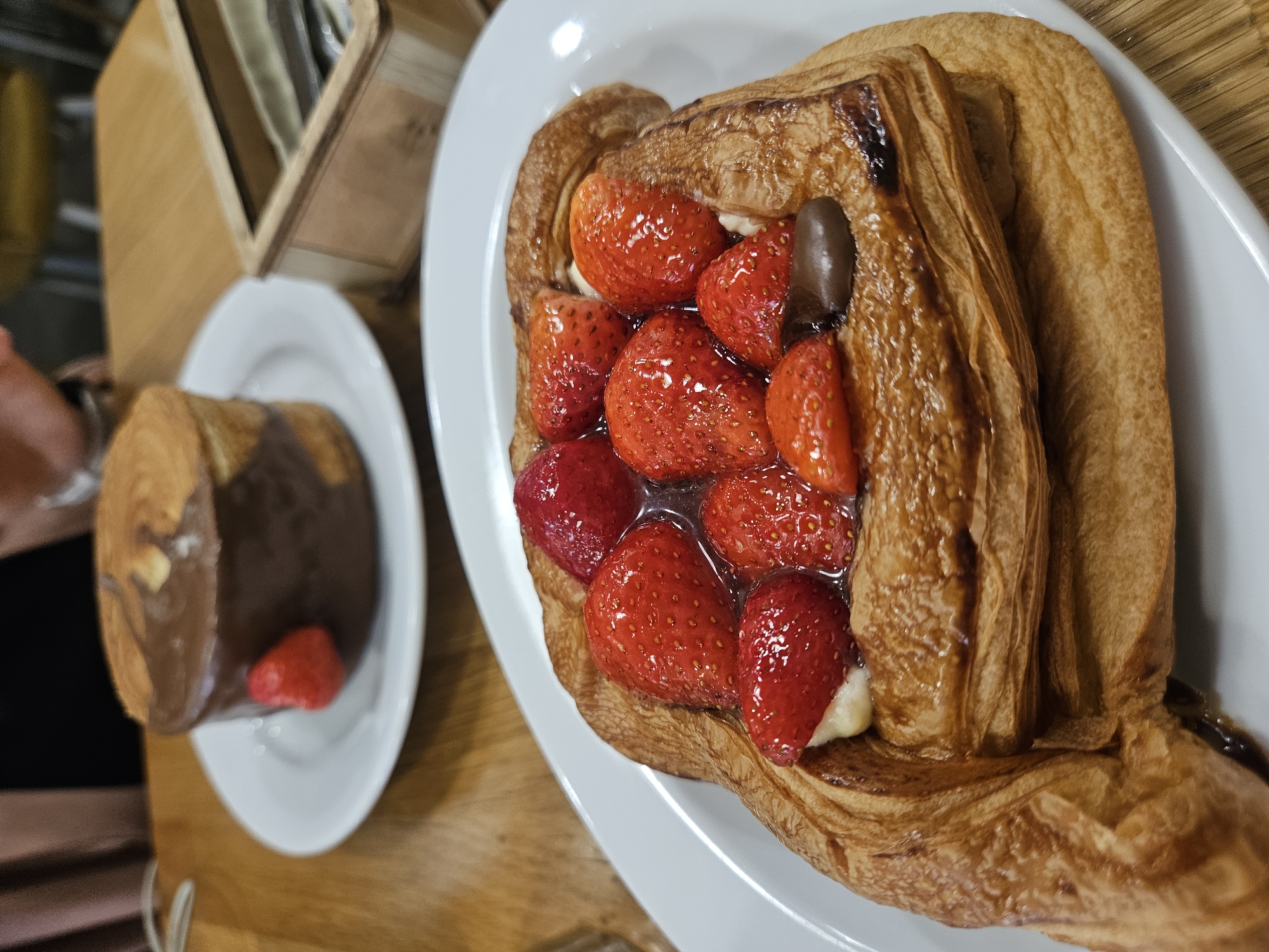 The image displays an overhead, slightly angled view of two pastries served on white plates atop a wooden table. The primary subject is a large, golden-brown laminated pastry, likely a Danish or similar, centrally placed on an oval white plate in the foreground. This pastry is generously filled with numerous fresh, bright red strawberries, many of which appear glossy, suggesting they are glazed or in a light syrup. Beneath the strawberries, a white cream or custard is visible. A single dark, elongated piece of chocolate or a thick chocolate drizzle is also present among the fruit.
In the blurred background, to the left, a second, smaller white plate holds another pastry. This pastry is mostly covered in a rich, dark brown chocolate coating and appears to be standing upright. A single, smaller red strawberry rests at its base.
The setting is an indoor environment, characterized by a warm wooden table surface. A light-colored wooden caddy or holder, possibly for napkins or utensils, is partially visible in the upper left background. A hand or arm is also partially visible on the far left. The lighting is bright, indicating it is likely daytime. There is no visible text in the image. The scene evokes a feeling of indulgence and freshly prepared treats.