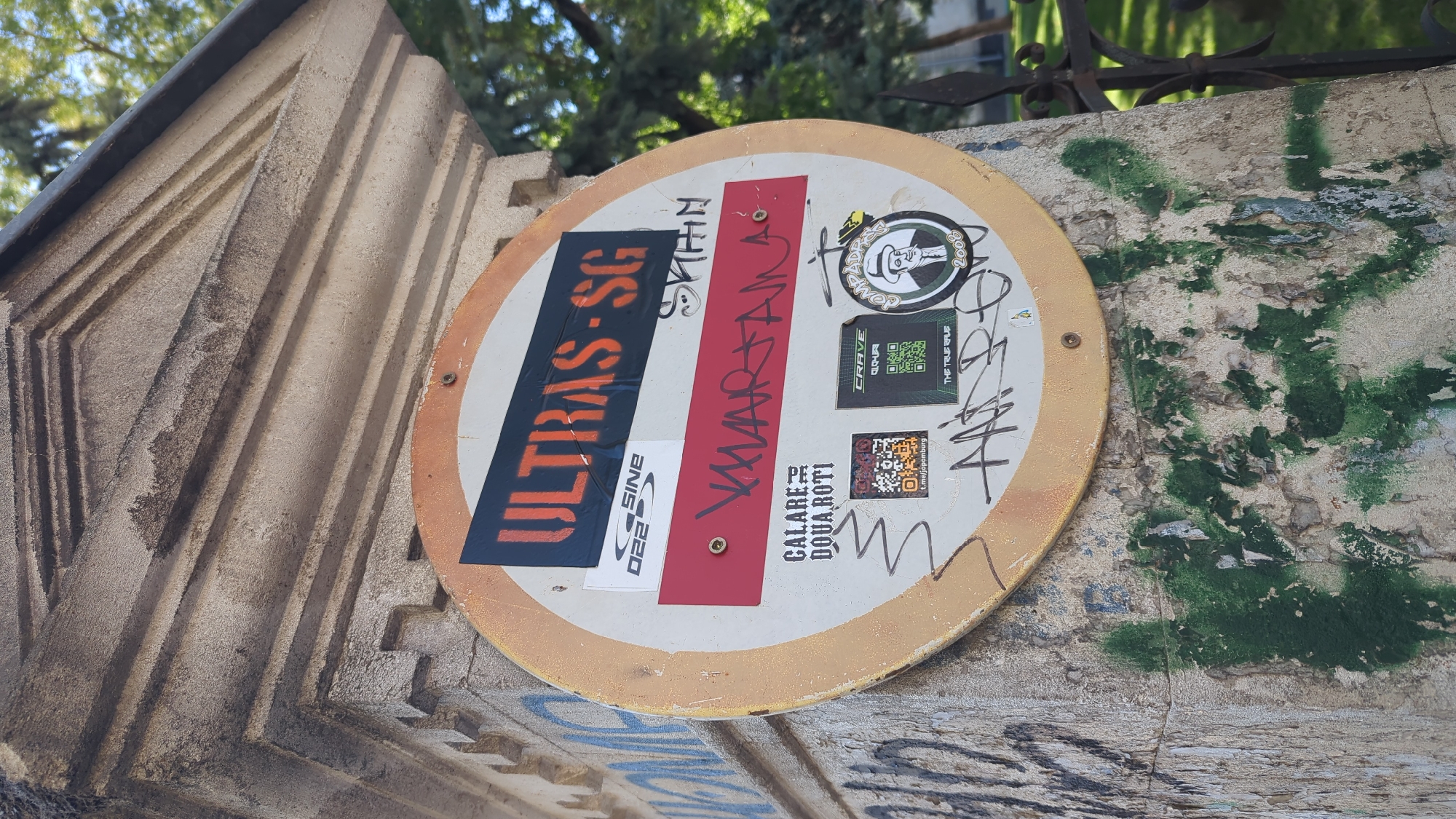 The image shows a round sign on a stone pillar, possibly part of an entrance or monument. The sign is covered with stickers, some of which contain text and graphics. One prominent sticker has "ULTRAS · SG" in orange spray paint on a black background. Below that, there's a red sticker with stylized red lettering that appears to say "VARTAN". Several other smaller stickers are present, including QR codes and logos. The pillar itself is weathered and shows signs of graffiti and peeling paint. The background includes trees and a metal fence, suggesting an outdoor urban environment.
Without further context or recognizable landmarks within the image, it is impossible to pinpoint the exact location within Chișinău, Moldova.
