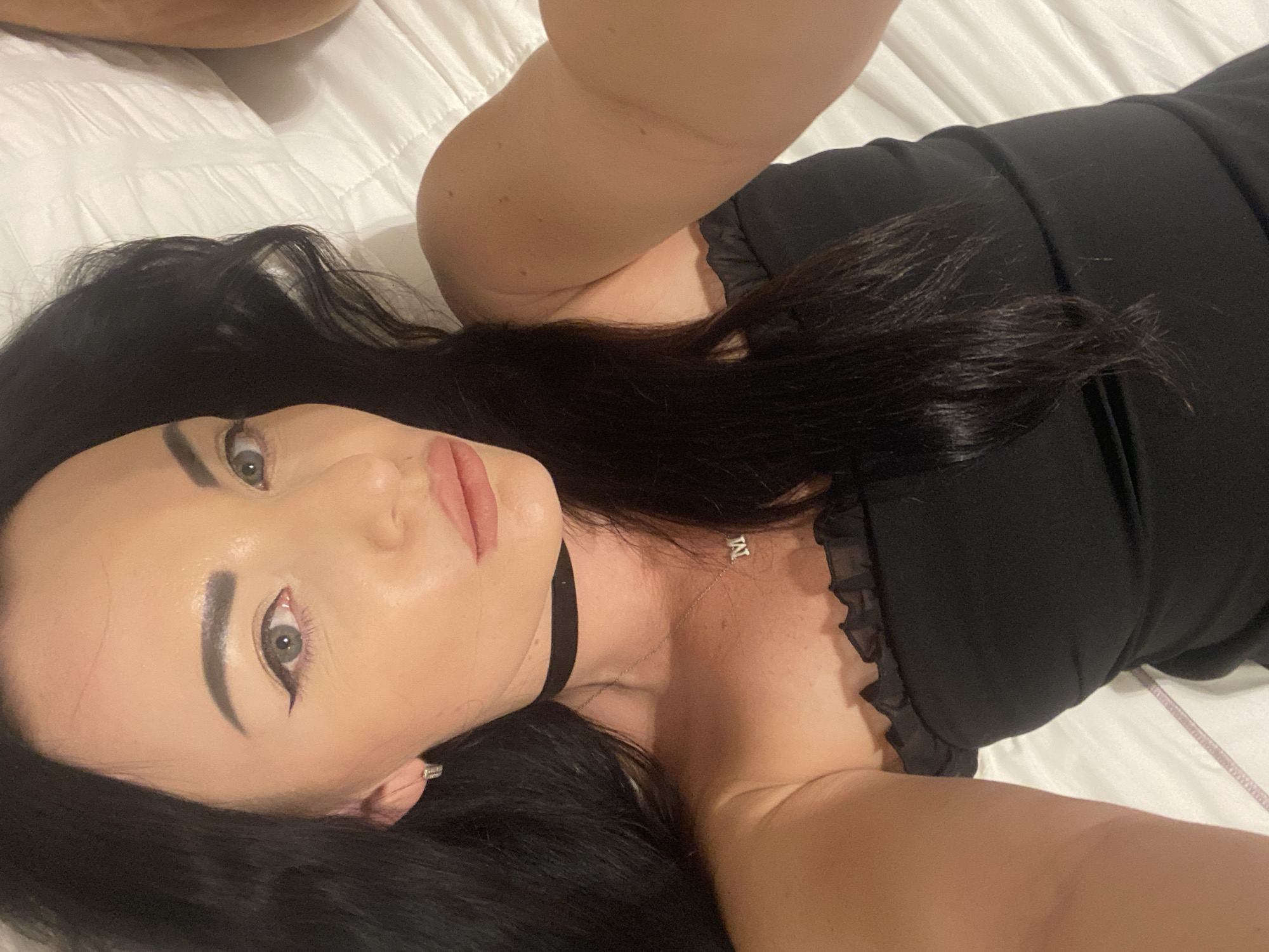 A woman with long, dark hair and fair skin lies on a white, quilted surface, possibly a bed. She is looking at the camera with a neutral expression. She is wearing a black strapless top with a ruffled neckline, a black choker necklace, and a silver necklace with a pendant shaped like the letter "M". Her makeup includes winged eyeliner and light lipstick. Her left arm is raised, holding a camera, suggesting she is taking a selfie. The background is a white, quilted surface, possibly a bed or couch. No specific landmarks of Sharm El Sheikh, Egypt are visible in the image.