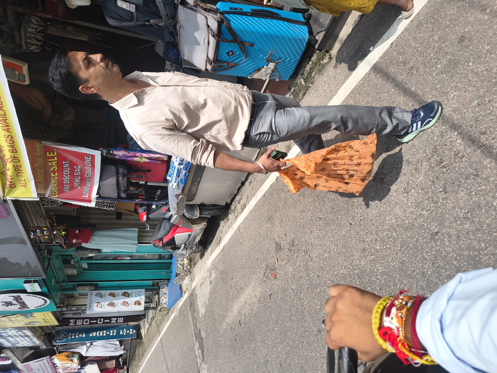 This image captures a street scene in Dharamshala, India, likely in the bustling market area. A man in a light-colored shirt and gray pants walks across the asphalt road, carrying an orange shopping bag in his right hand. He is wearing a watch on his left wrist and dark sneakers with light green accents. To his right, a large, bright blue suitcase stands upright next to a shop entrance. In the foreground, a person's hand with several colorful bracelets is visible, holding onto a handlebar, suggesting the image is taken from a moving vehicle or bicycle. The background features shops lining the street, displaying clothing, bags, and other merchandise. A red scooter is parked on the sidewalk. The overall atmosphere is that of a typical Indian marketplace on a sunny day.
