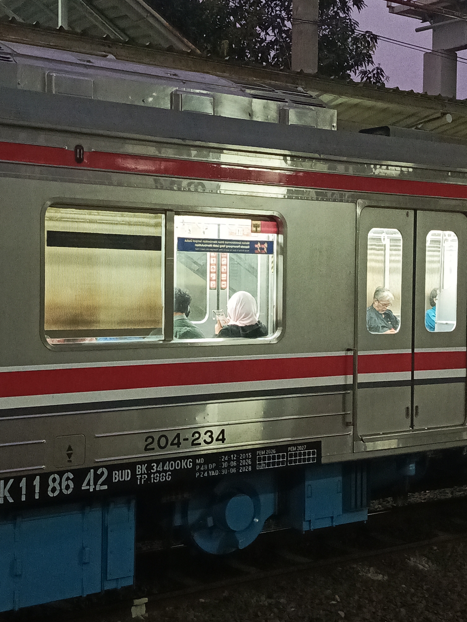 This image depicts the exterior of a train car at a station in Telagamurni, Indonesia. The train is silver with a red stripe running horizontally along its side. There are several windows visible, some with people inside. In one window, two individuals are seated, with one wearing a headscarf and appearing to look at a mobile device. Another window shows an older person with glasses, reading a book. A third window has a person in a blue shirt. The train car bears several markings, including the number "204-234" and "K1186 42". The environment appears to be dusk or early evening, with a dusky sky visible above the train and overhead structures. The train is positioned on tracks, with ballast visible beneath.