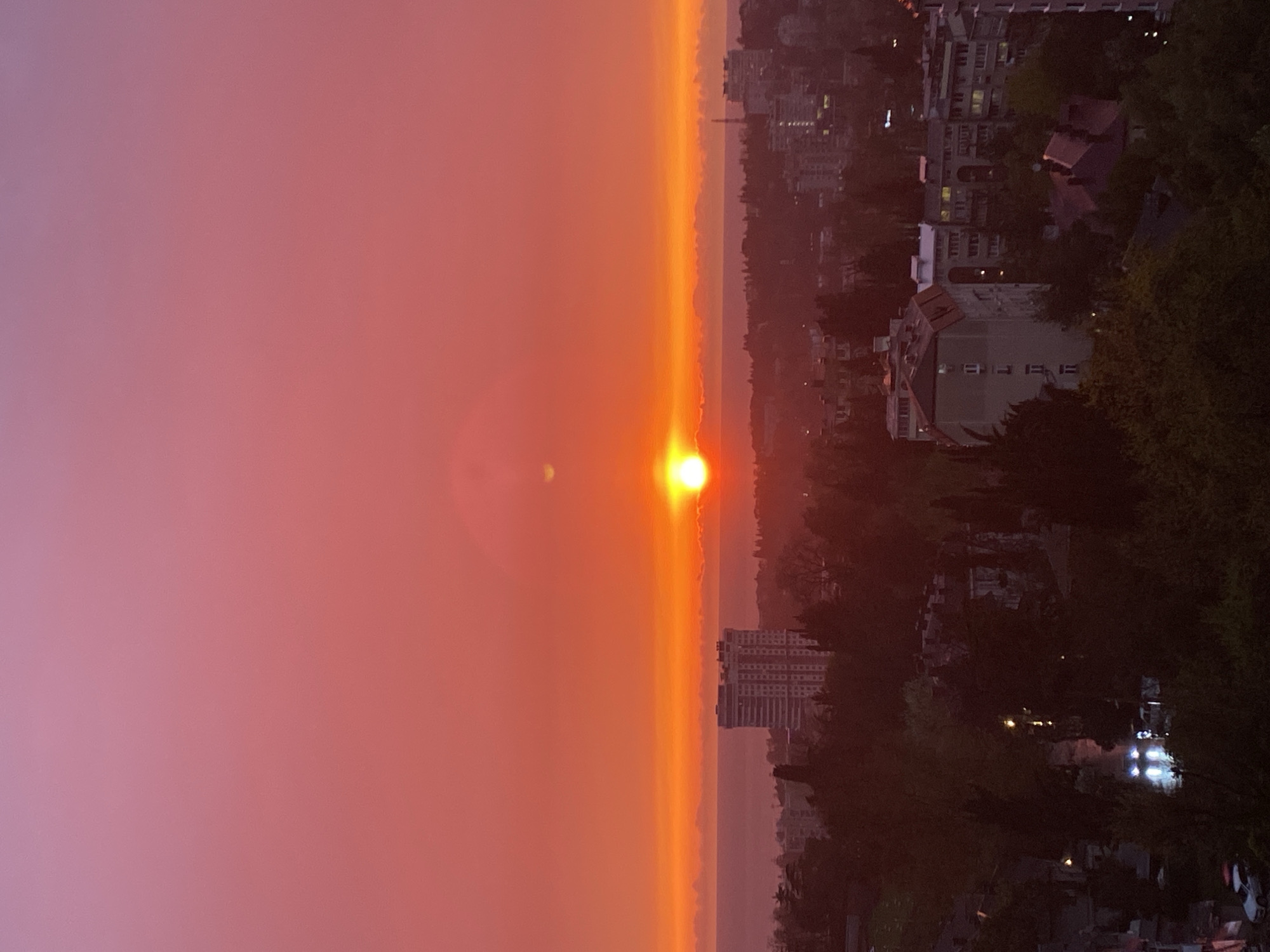 The image shows a sunset in Sochi, Russia. The sun is setting over the Black Sea, which is visible as a thin strip in the center of the image. The sky is a mix of orange, pink, and purple hues. On the right side of the image is a cityscape with buildings and trees. In the foreground, there are some dark, indistinct shapes that may be trees or bushes. The overall impression is one of a serene and beautiful sunset.