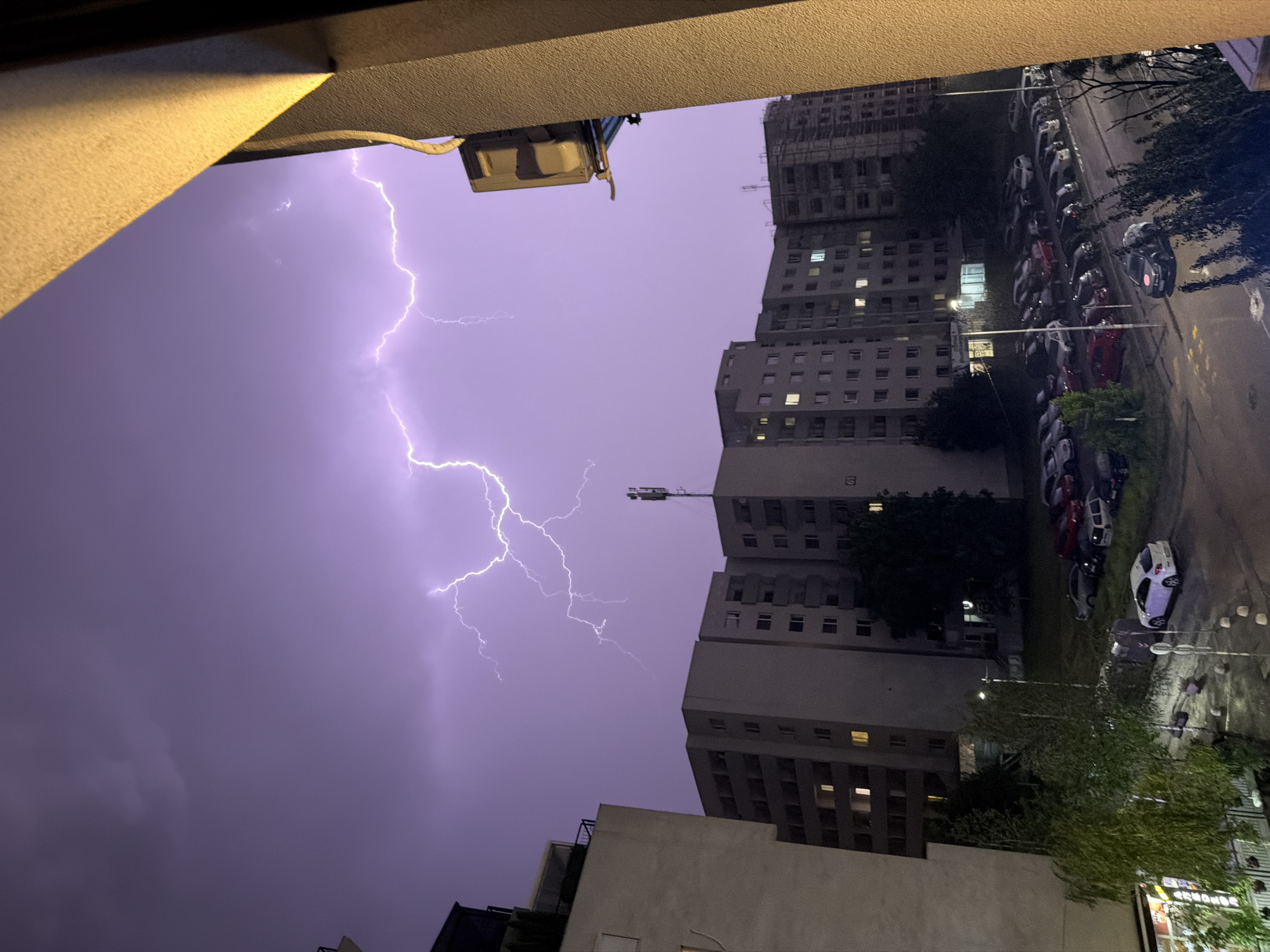 The image depicts a stormy night in Sofia, Bulgaria, as evidenced by the lightning illuminating the purple sky above apartment buildings. In the foreground, a balcony railing casts a shadow, suggesting the vantage point is from a residential building. The street below is lined with parked cars, some of which are illuminated by streetlights. The overall scene captures the dramatic and somewhat eerie atmosphere of a thunderstorm in an urban setting.