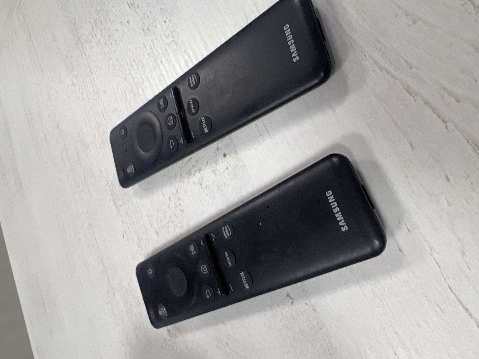 Two black Samsung TV remote controls are placed on a white surface with a wood grain pattern. The remotes are oriented in slightly different directions, with the button side facing upward. The "Samsung" logo is visible on both remotes. The location in Poltava, Ukraine is not discernable from the image.