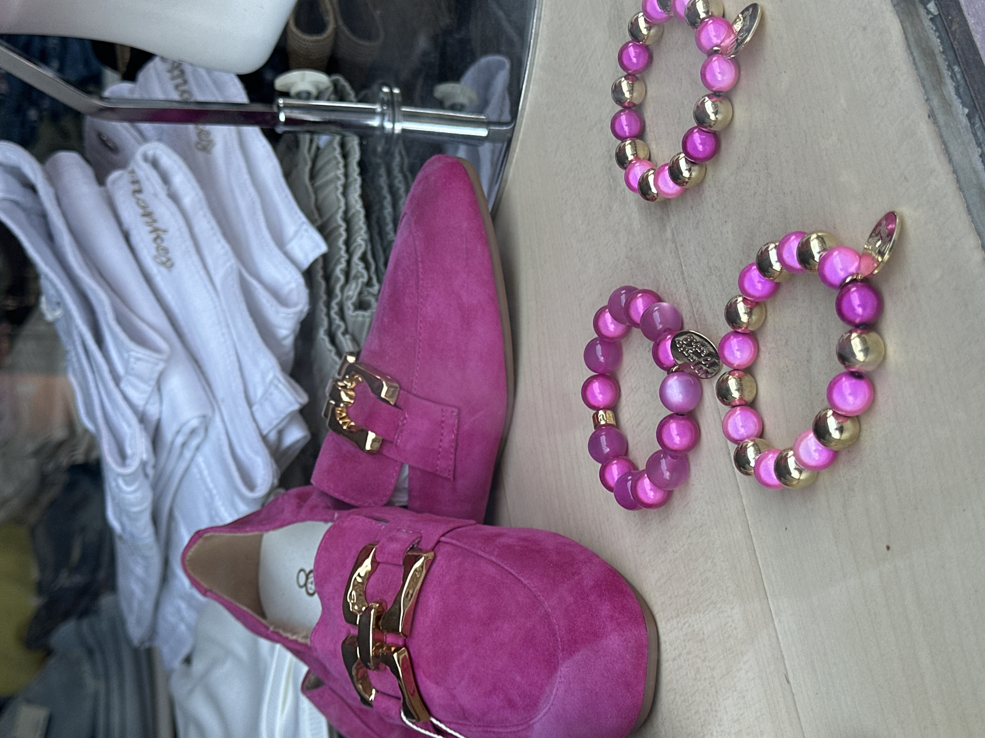 The image displays a pair of bright pink suede loafers with gold buckle detailing, positioned on a light-colored wooden surface. To the right of the shoes, two beaded bracelets, one above the other, are visible. These bracelets are made of alternating pink and gold beads. The background suggests a retail environment, with glimpses of white folded clothing, possibly t-shirts or blouses, on a rack and hangers. The overall lighting appears to be natural, possibly from a storefront display. There are no people or specific weather conditions indicated. The visible text on the shoe's insole reads "ogo."