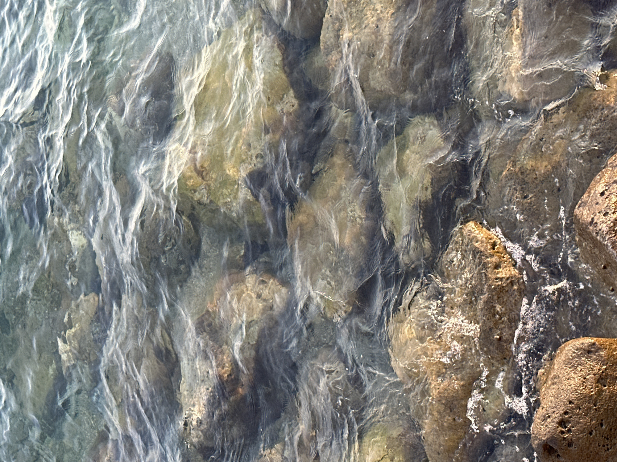 The image shows a close-up view of clear, shallow water flowing over submerged rocks. The rocks are varied in size and color, ranging from dark grey to reddish-brown, with some appearing smooth and rounded, while others are rough and porous. Sunlight penetrates the water, illuminating the underwater landscape and creating shimmering reflections on the surface. Small ripples and waves are visible, indicating gentle movement of the water. The scene appears to be a natural coastal environment, possibly along the shoreline of Budva, Montenegro, given the rocky, aquatic setting. There are no people or man-made objects depicted in the image.