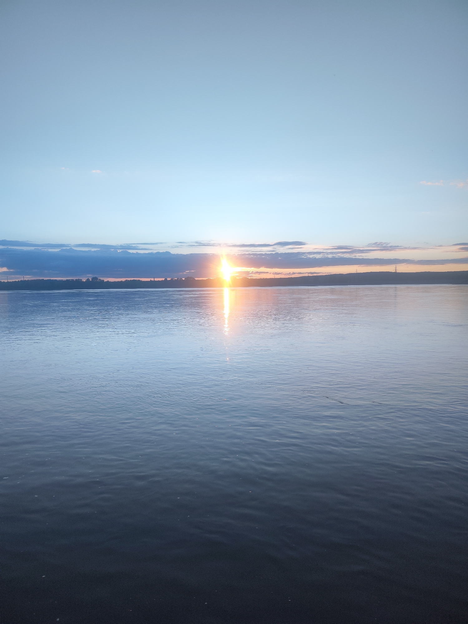 This image presents a serene sunrise or sunset over a vast body of water, likely a river or a large lake, near Noviy, Russia. The sun, a brilliant point of light, is positioned low on the horizon, slightly to the left of the frame's center, casting an intense, shimmering vertical reflection that stretches across the water towards the viewer.
The sky above is a gradient of soft blues, transitioning to warmer orange and pink tones closer to the horizon where the sun is. Scattered clouds, some dark and elongated, are visible, particularly above and around the sun, adding texture to the sky. A distant landmass or shoreline appears as a low, dark silhouette against the vibrant sky, with faint outlines of what seem to be man-made structures, possibly power line towers, visible along its length.
The water surface is calm with gentle ripples, reflecting the subdued light of the sky and the intense gleam of the sun. The foreground water appears deeper blue and subtly textured by the ripples. No people or specific activities are visible in the tranquil scene, which emphasizes the natural landscape. The time of day is clearly dawn or dusk, characterized by the low sun and soft, transitioning light. No visible text is present in the image.