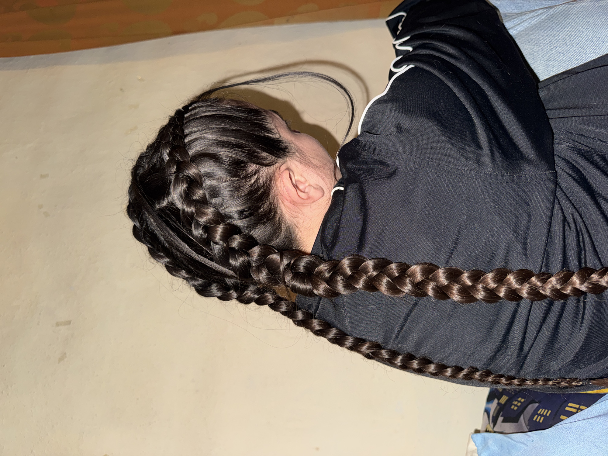 The image shows the back of a person with long, dark brown hair styled in two thick braids. The braids are tightly woven, and the hair appears healthy and shiny. The person is wearing a black athletic jacket with white trim. The background is a plain, textured wall, likely an interior wall of a building. There is also a hint of a window or curtain on the right side of the image with an orange hue. The lighting suggests an indoor environment, possibly in the evening or a dimly lit room. The scene does not provide any specific indicators of its location within the city of Ruzhitsa, Bulgaria.