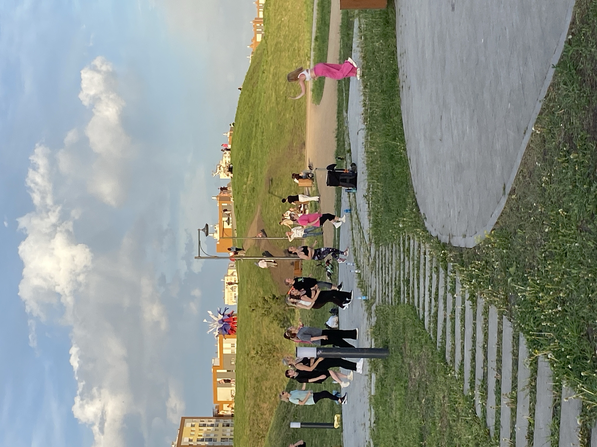 Here is a description of the image:
The image shows a group of people in a park, likely Gorky Park in Yekaterinburg, Russia. There is a grassy hill to the left with people sitting or lying on it. A path runs along the base of the hill. Closer to the viewer is a walking path constructed with stone pavers and sections of concrete. A group of people are standing and interacting with one another near the walking path, suggesting a gathering or exercise class. A young girl is jumping in the air, and other people are seated along the path. In the background, there are buildings and a sky with some clouds.