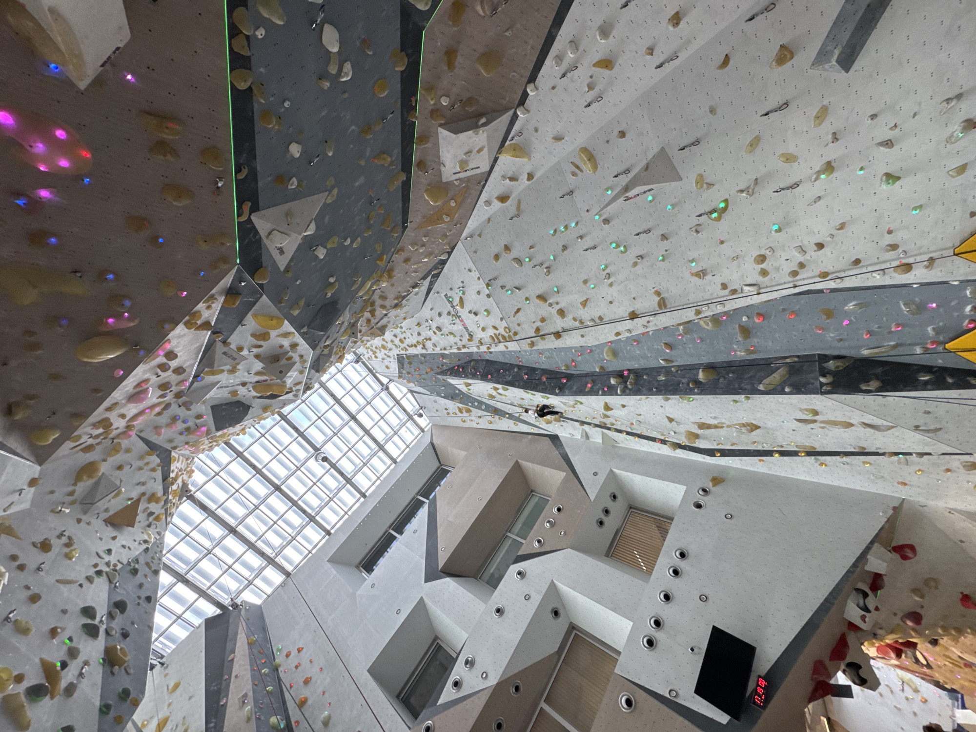The image shows the interior of a climbing gym in an unknown city. The walls are covered in climbing holds of various shapes and colors. The walls are gray and white in color. The room is well-lit, with a large skylight overhead. Windows on the side of the wall with the time display. There are no people visible in the image. A digital clock is visible, displaying "17 19 09."