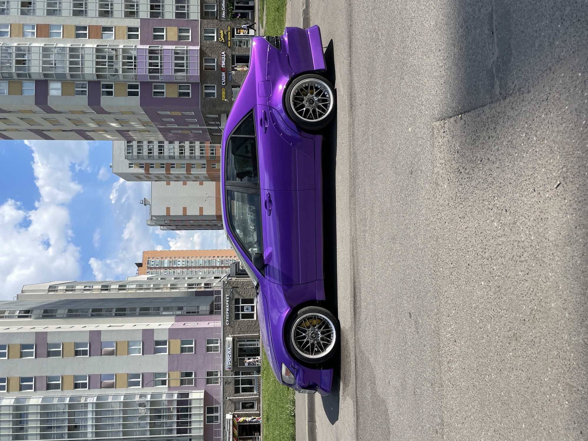 A customized, low-slung, four-door sedan, painted a vibrant metallic purple, is parked parallel to the curb on an asphalt road in a residential area of an unknown city in Russia. The car, consistent with a first-generation Lexus IS or Toyota Altezza, features prominent riveted wide-body fender flares, aftermarket front and rear bumper treatments, and multi-spoke polished alloy wheels with visible yellow brake calipers.
The background consists of several multi-story modern apartment buildings, primarily colored light gray with contrasting horizontal and vertical bands of purple and beige. The ground floors of these buildings host commercial establishments. On the left, a store labeled "РОСАЛ" and "СУПЕРМАРКЕТ" (SUPERMARKET) is visible. On the right, signs for "ЛИНВО," "СУШИ ПИЦЦА" (SUSHI PIZZA), "Smoke," and "Alex.Coffee" are legible. Taller, orange-brown buildings are visible in the distant background.
A narrow strip of mown green grass separates the road from the sidewalk. Several casually dressed individuals are visible on the sidewalks: two near the "РОСАЛ" entrance, and one walking near "Alex.Coffee." The scene is illuminated by bright sunlight under a clear blue sky with scattered cumulus clouds, indicating fair daytime weather. Shadows from the car and buildings are distinctly cast on the asphalt road, which shows minor wear and a few faint patches.