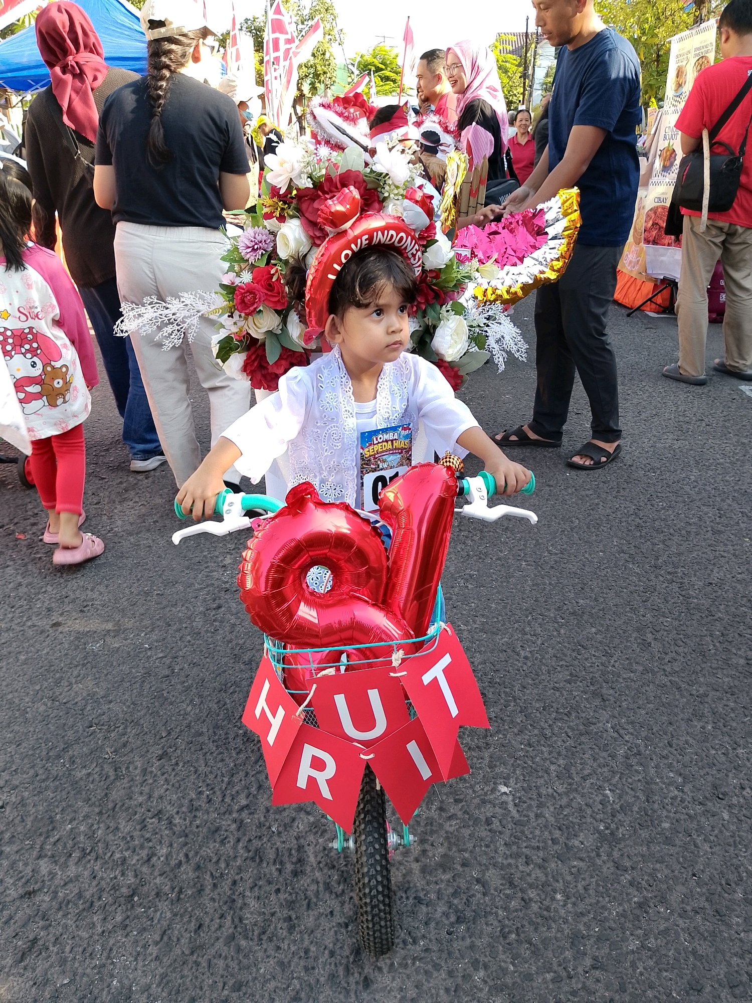 This image depicts a young girl in the foreground, sitting on a bicycle decorated with red balloons and a banner that reads "HURT". The bicycle also has a bouquet of flowers attached to its front. The girl is wearing a white traditional Indonesian outfit and looking to the right. She is participating in a "Lomba Sepeda Hias" (Decorated Bicycle Competition) in Surabaya, Indonesia, as indicated by the event's signage on her chest. In the background, several adults and children are visible, suggesting a festive street event. The setting appears to be an urban environment, with asphalt pavement and onlookers gathered around. The overall atmosphere is one of celebration and community.