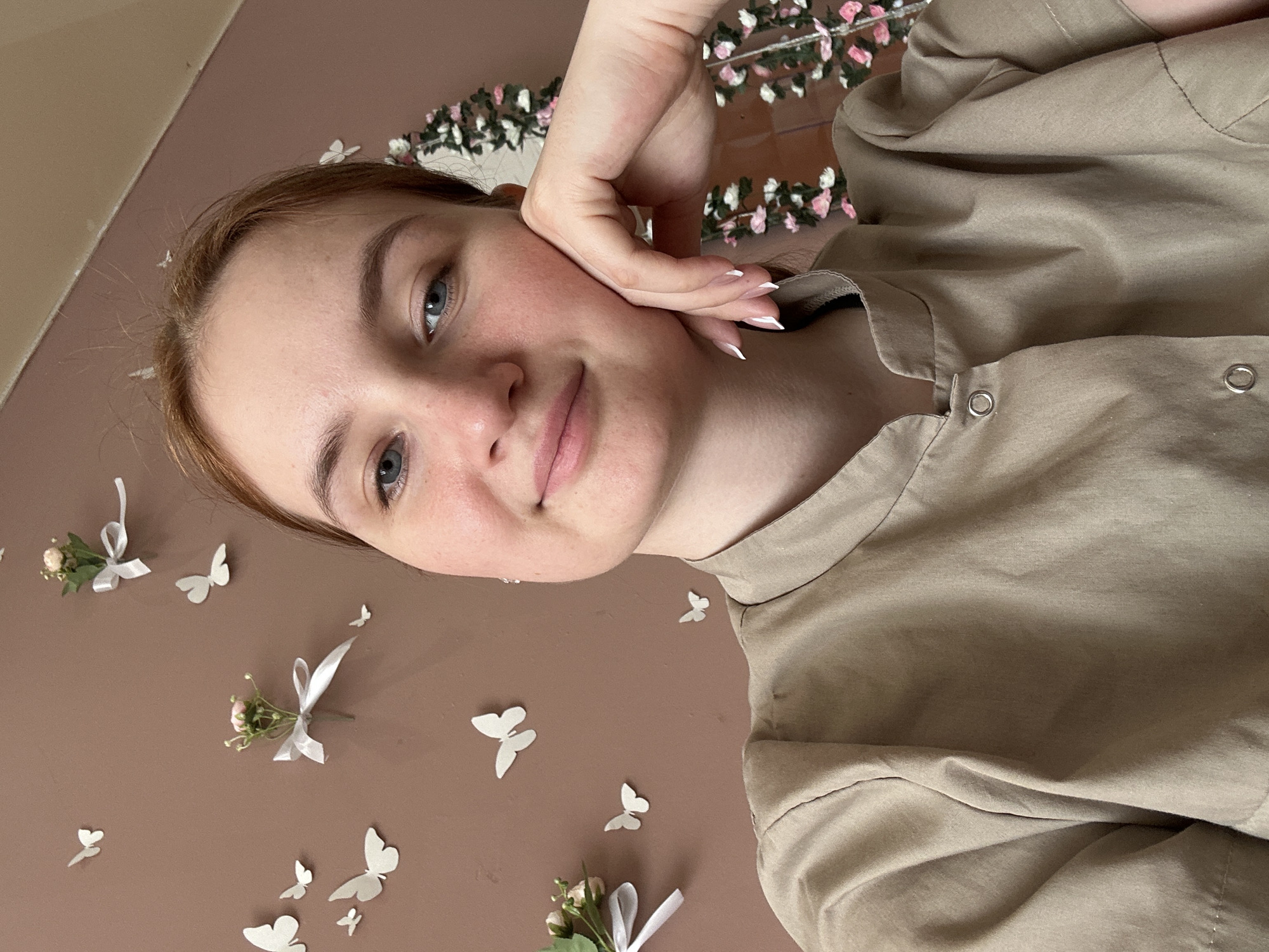 A young woman with light skin and auburn hair poses for a selfie. Her left hand is resting on her cheek. She is wearing a tan collared shirt with button details. The background features a light brown wall decorated with small white paper butterflies and miniature floral arrangements tied with white ribbons. A floral garland with pink and white flowers is visible in the upper background. The photograph appears to have been taken in an indoor setting. It's unknown if the location is Chaikovsky, Russia.