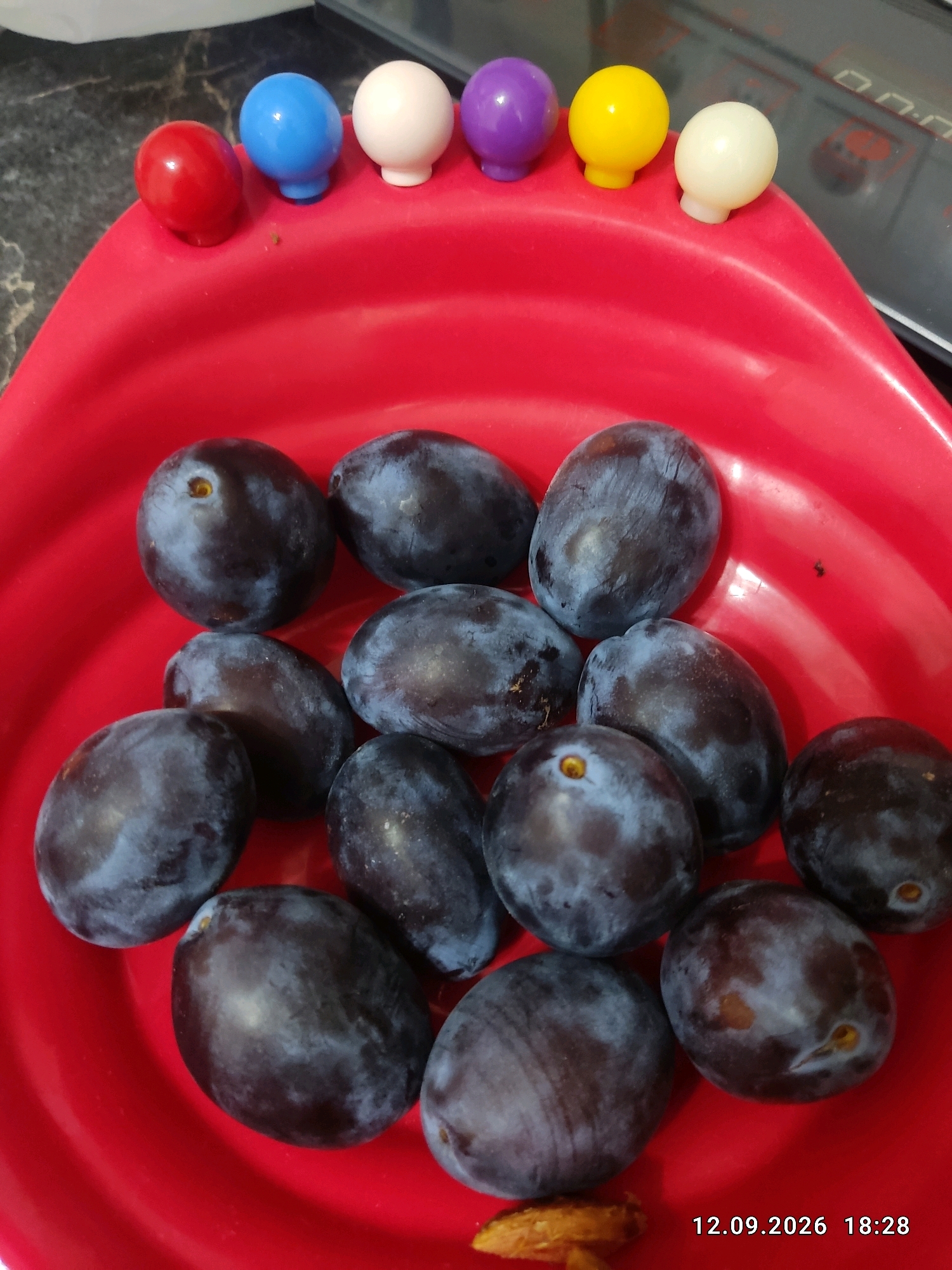 A top-down, slightly angled shot shows a bright red, plastic bowl filled with ripe plums. The plums are dark purple, some with a bluish bloom, and vary in shape from oval to slightly round. They are clustered together in the bowl. At the top edge of the bowl, several small, brightly colored plastic spheres are visible: red, blue, white, purple, yellow, and another white sphere. A sliver of a brown, dried fruit is visible at the bottom of the bowl. The background is a dark, textured surface, likely a countertop. A digital timestamp, "12.09.2026 18:28", is overlaid in the bottom right corner. The image does not contain information about Bishkek City, Kyrgyzstan.