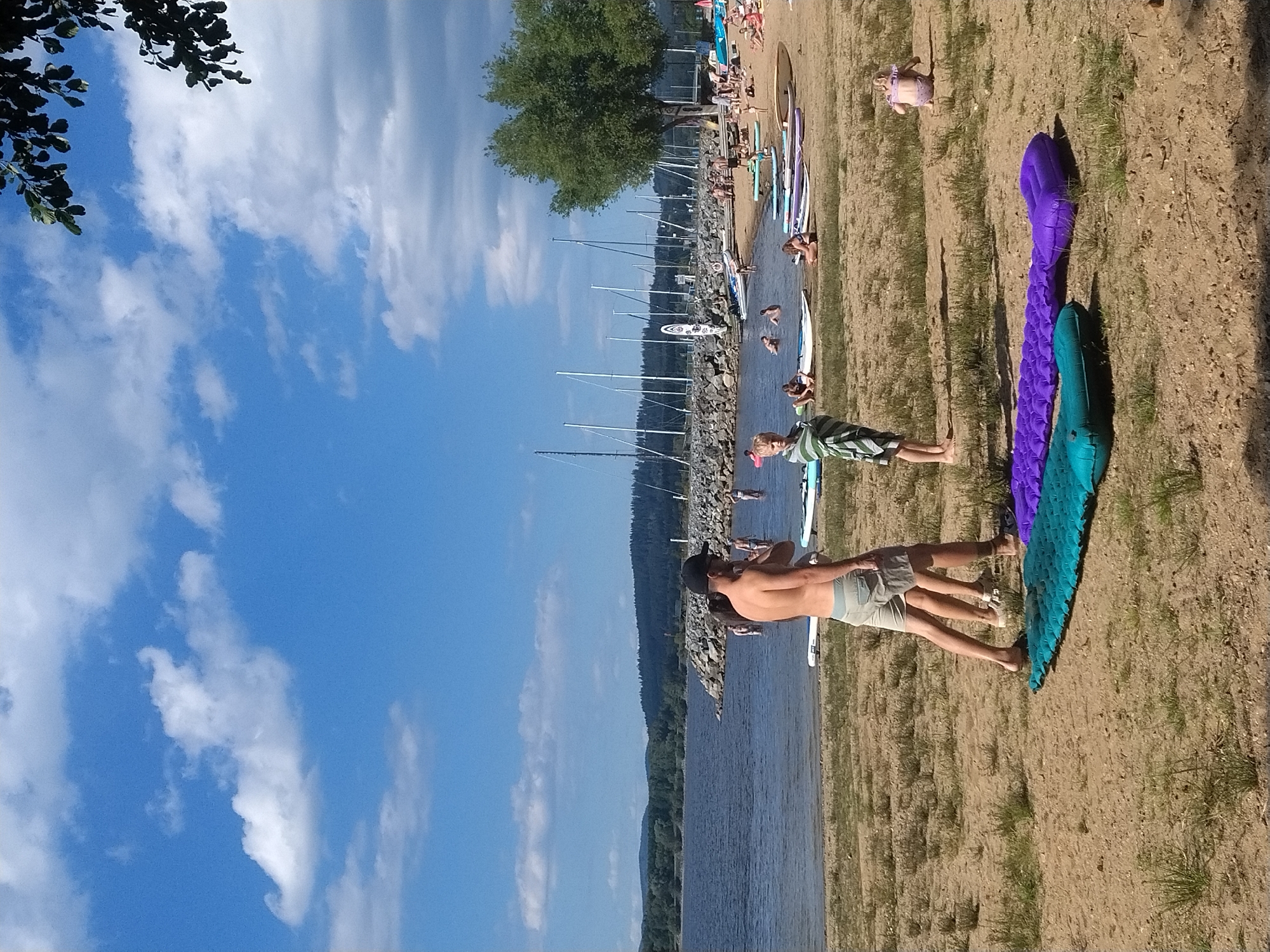 The image depicts a sunny day at a lakeside beach in Radslav, Czechia. In the foreground, two inflatable mattresses, one turquoise and one purple, lie on the sandy ground. Several people are visible near the water's edge and on the beach. A man wearing a black cap and shorts stands with his back to the camera. A child wrapped in a striped towel stands to his left, facing the viewer. Further back, various individuals are swimming or wading in the water, and some are relaxing on the sandy shore. Paddleboards and other watercraft are lined up on the beach and in the shallow water. In the background, a rocky breakwater juts into the lake, and beyond that, a dense forest covers rolling hills. Numerous sailboat masts are visible on the water, suggesting a marina or sailing club. The sky is blue with scattered white clouds.