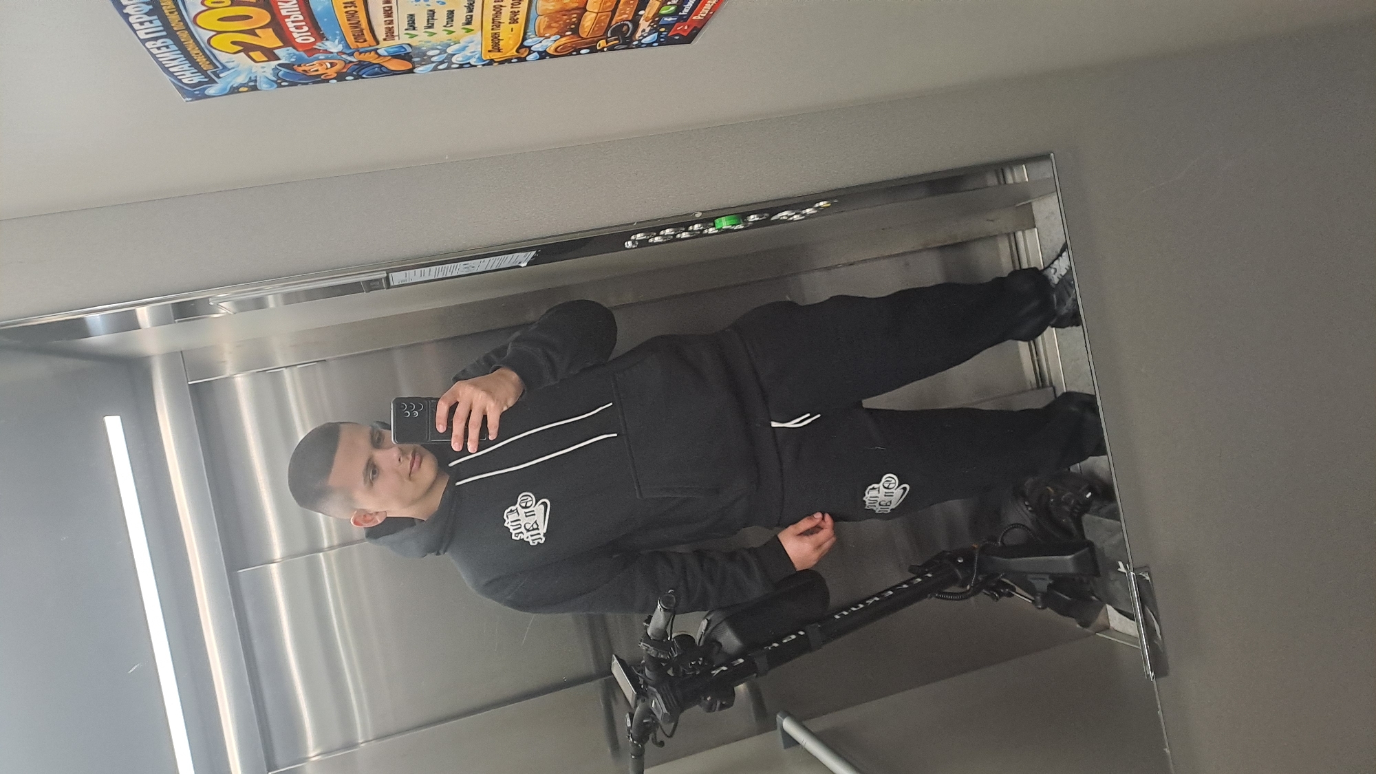 A man in a black tracksuit and black shoes takes a selfie in an elevator. He is also holding an electric scooter. The elevator has metal walls and a row of green buttons. A poster advertising cleaning services is visible near the ceiling, indicating the location is Burgas, Bulgaria. The poster has text in Bulgarian and references "SHANIKOV PERFO" and "20% OTS'TPKA".