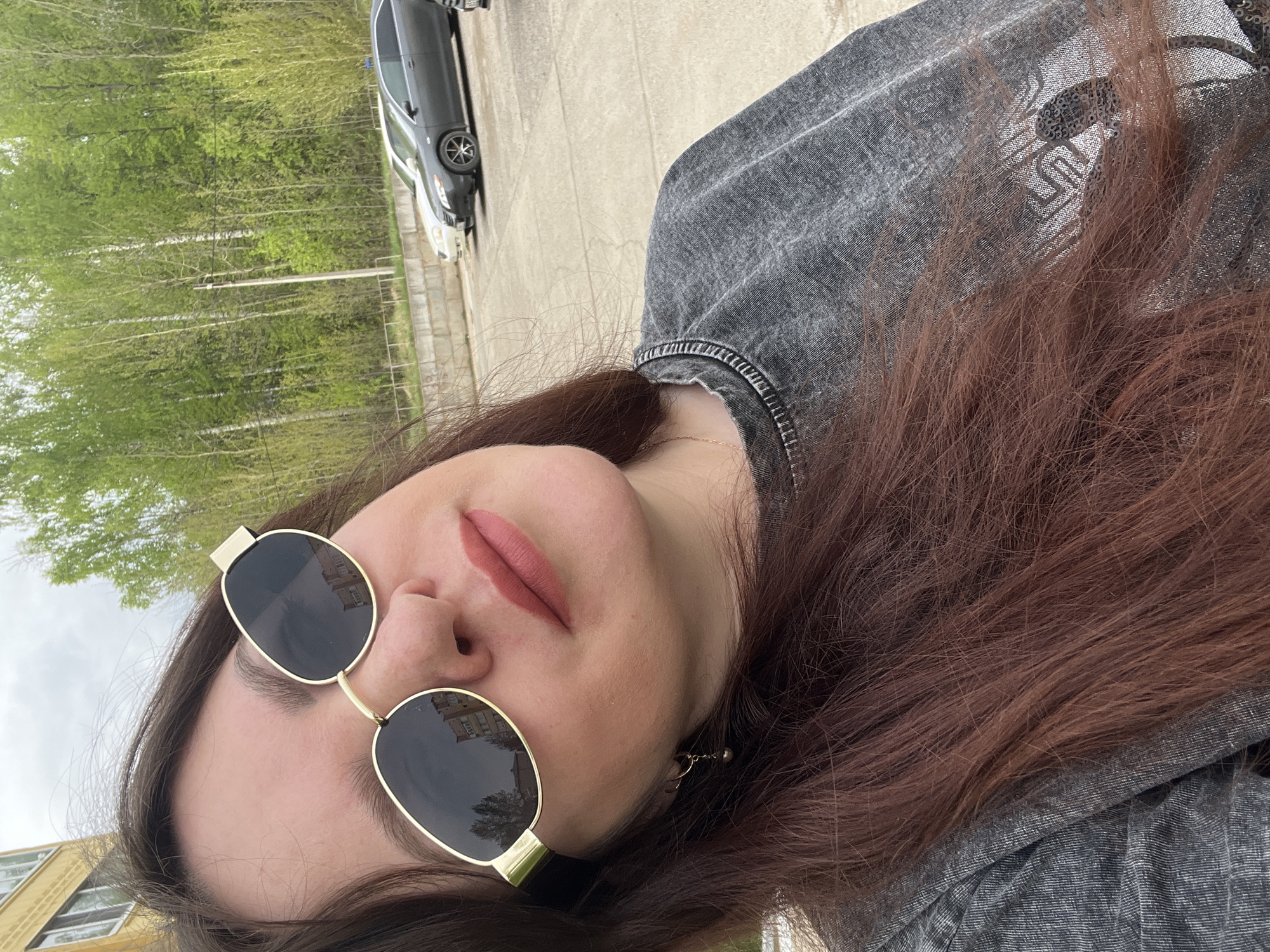 A woman with auburn hair and wearing sunglasses is posing for a photo in an outdoor setting. The background features trees and a gray car parked on concrete. The reflection in the sunglasses shows a building. The location is likely Noviy, Russia, based on previous context. The woman is wearing a gray denim jacket.