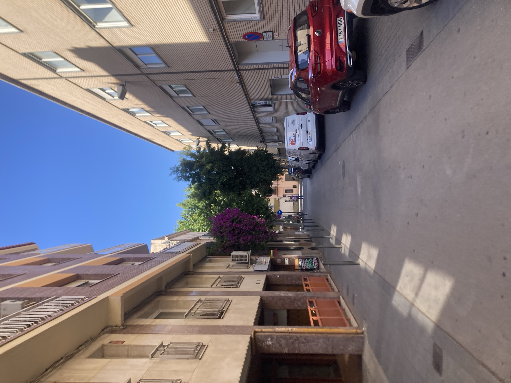 The image depicts a narrow urban street in Badalona, Spain, flanked by multi-story buildings under a clear blue sky. On the left, older residential or mixed-use buildings feature balconies with white railings, exposed conduits, and ground-level storefronts. One building on the left has an air conditioning unit mounted below a dense patch of vibrant purple bougainvillea-like flowers. A menu board is visible outside one ground-floor establishment. Bollards line the left sidewalk, casting distinct rectangular shadows onto the pavement.
On the right, a more uniformly beige brick building with multiple windows and attached streetlights extends along the street. Several vehicles are parked parallel to the curb on the right side. These include a white commercial van with "INSTAL LADUR" text and Spanish license plate "0386 MNK", followed by a red MG ZS SUV with Spanish license plate "7161 NBN". The front of another white vehicle is partially visible in the bottom right corner. A red and blue "no parking" sign is affixed to the building on the right.
Further down the street, at its visible terminus, an intersection is faintly discernible. Sparse trees are present, and a single pedestrian is observed walking towards the viewer in the distance. The street and sidewalks are paved, showing signs of wear, and bright sunlight creates sharp shadows from the buildings and bollards. The overall scene suggests a quiet, sunny day in a residential or light commercial urban environment.
