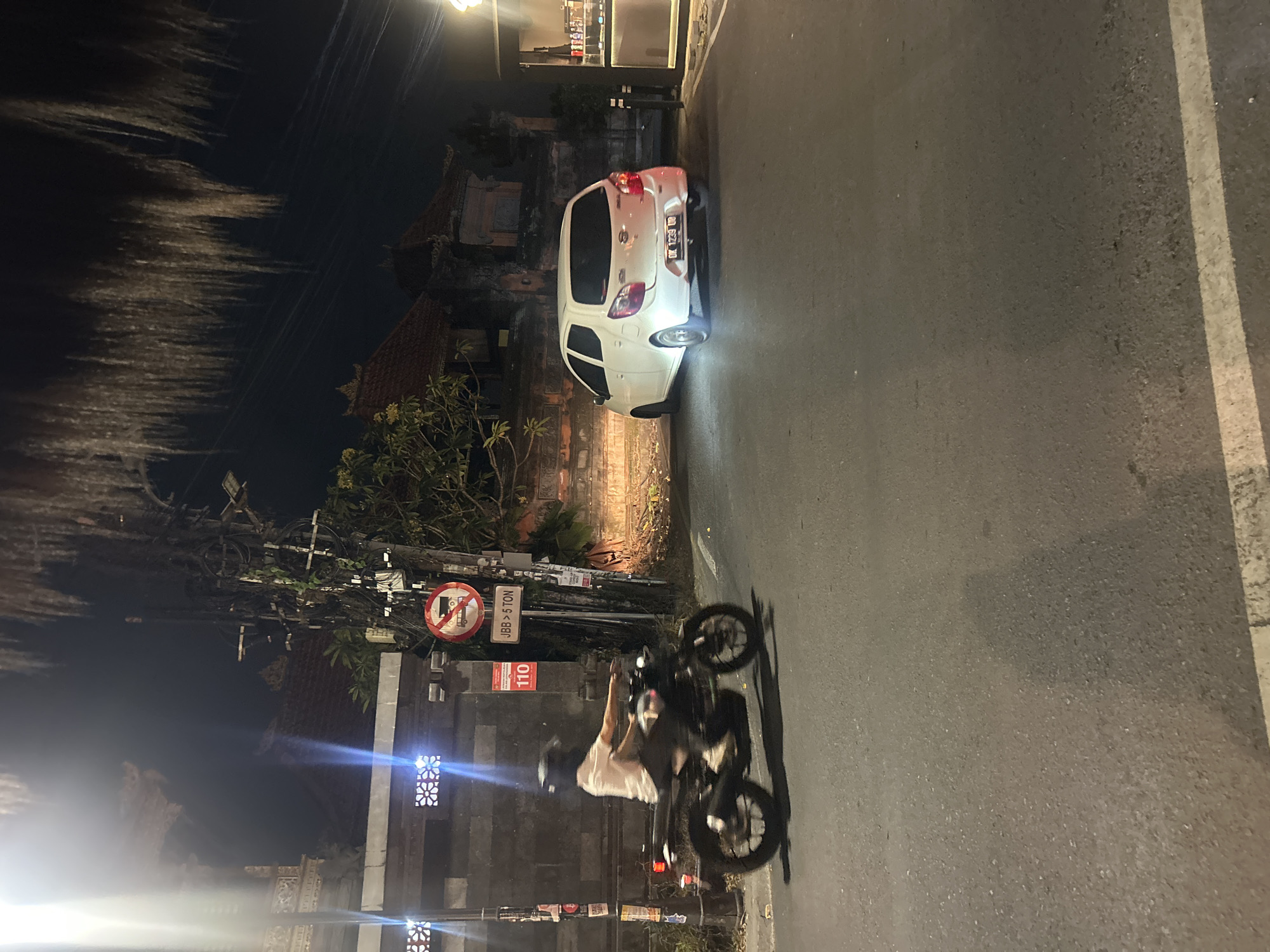A nighttime street scene in North Kuta, Indonesia, captures a white car stopped on the right side of the road, its headlights illuminating the asphalt. To the left, a person on a motorcycle is in motion, their form blurred by the camera's exposure. The surrounding environment features a stone wall with decorative elements, a utility pole adorned with numerous wires and signs, and traditional Indonesian architecture with tiled roofs in the background. Overhead, palm fronds hang, adding to the tropical ambiance. The street itself is paved, with a white line marking the edge of the lane.