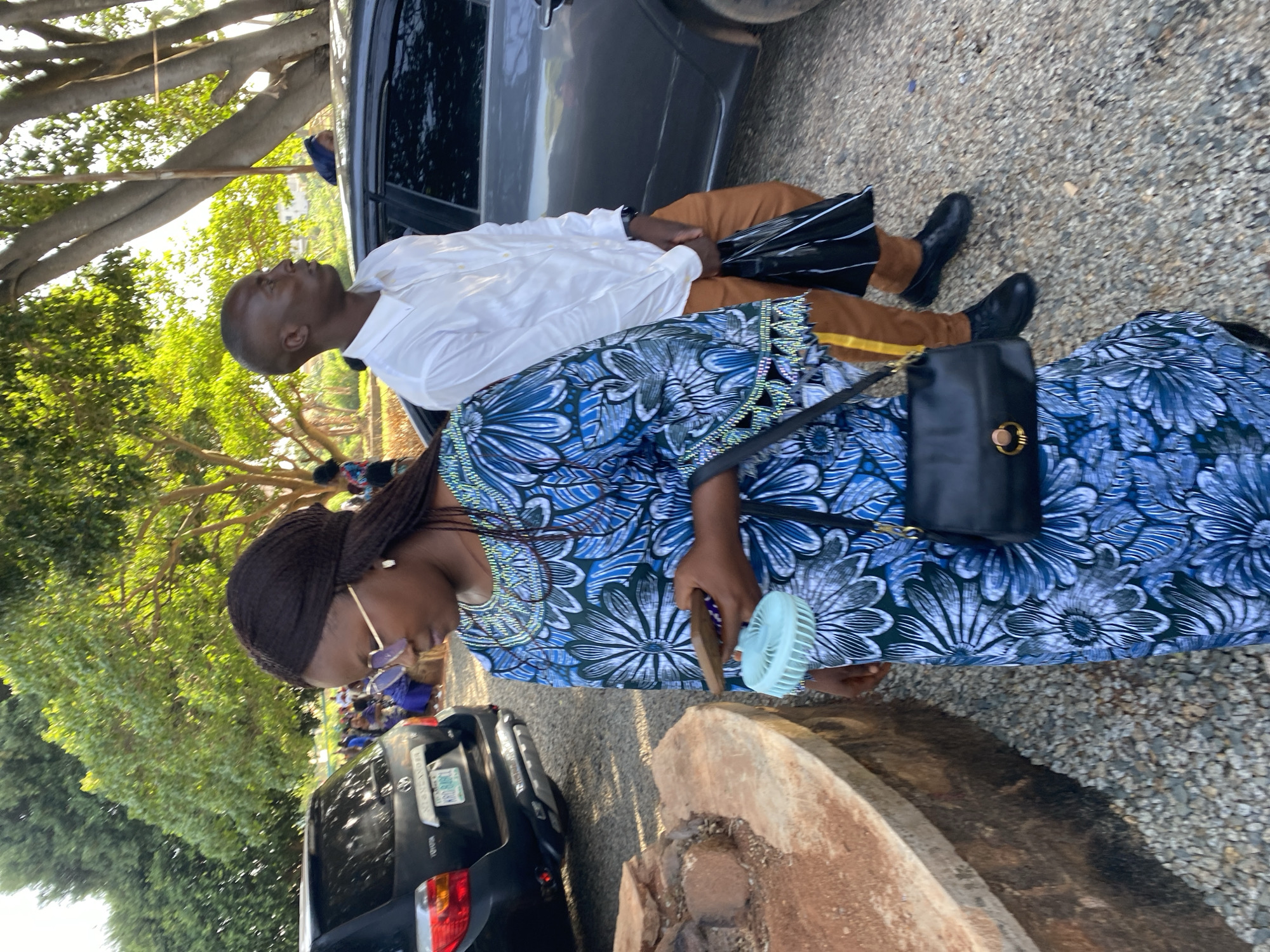 The image depicts two individuals standing near parked vehicles on a gravel surface, likely outdoors. The first person, a woman, wears a blue patterned dress with a black purse and is holding a small fan. The second person, a man, wears a white shirt and brown trousers with black shoes. A black umbrella is seen near him. The scene takes place near cars with Nigerian license plates, and the location appears to be in Jos, Nigeria. The background includes trees and other people.