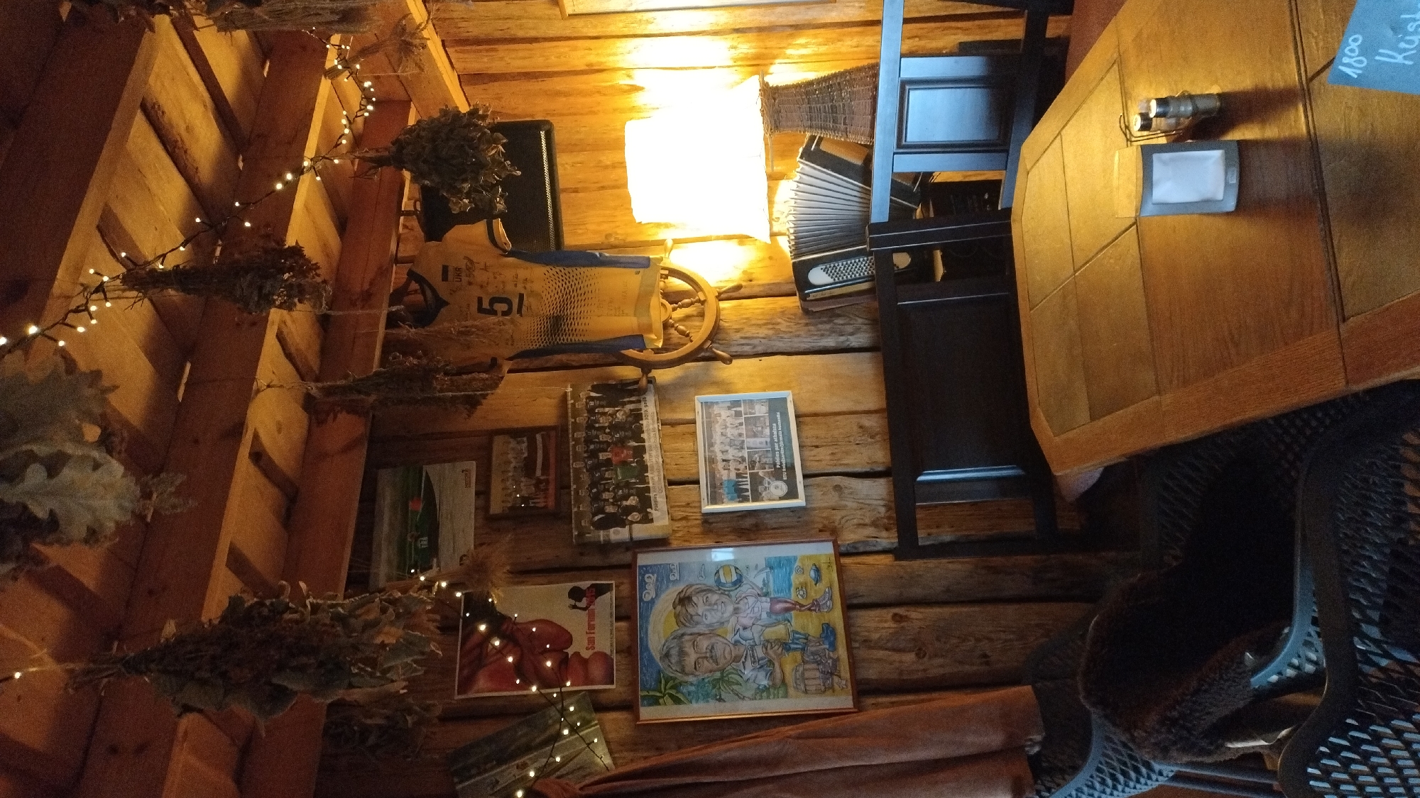 The image depicts the interior of a rustic establishment, likely a cafe or restaurant, characterized by wooden walls and ceiling. Dried herbs and foliage are hung from the ceiling, adorned with string lights. Several framed photographs and artwork are displayed on the walls, including a caricature of a couple, a group photo, and a poster. A sports jersey with the number "5" is also visible. On a wooden table in the foreground, there are salt and pepper shakers and a napkin holder. A lamp with a wicker shade is present, along with an accordion. The scene is lit by the warm glow of the lamp and string lights. The presence of the number "1800" and the word "Kuist" (possibly a name or a place) on a small sign in the lower right corner might indicate a location within Jūrmala, Latvia, however, this cannot be definitively confirmed without further context.