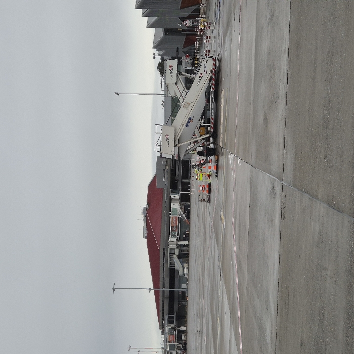The image displays an airport apron under an overcast, grey sky in Blagnac, France. The ground consists of wet asphalt or concrete, exhibiting visible expansion joints and horizontal red and white striped markings in the foreground.
In the mid-ground, a white self-propelled passenger stair unit, bearing the "TCR" logo and additional blue and red markings, is positioned facing left. The unit's stairs are elevated. It is cordoned off by several orange safety cones and white-and-red striped barriers. A solitary individual, dressed in high-visibility clothing, is standing near the lower left side of the stair unit.
The background features two primary structures. To the left, an airport terminal building with a distinctive reddish-brown, slightly peaked roof is visible. This building appears multi-level with numerous windows and several jet bridge structures extending from it. To the right, another building with a dark, angular, possibly corrugated or sawtooth-style metallic roof is present. Multiple tall, metallic light poles are distributed across the apron area. The overall scene suggests an operational airport environment during a cloudy, damp period.