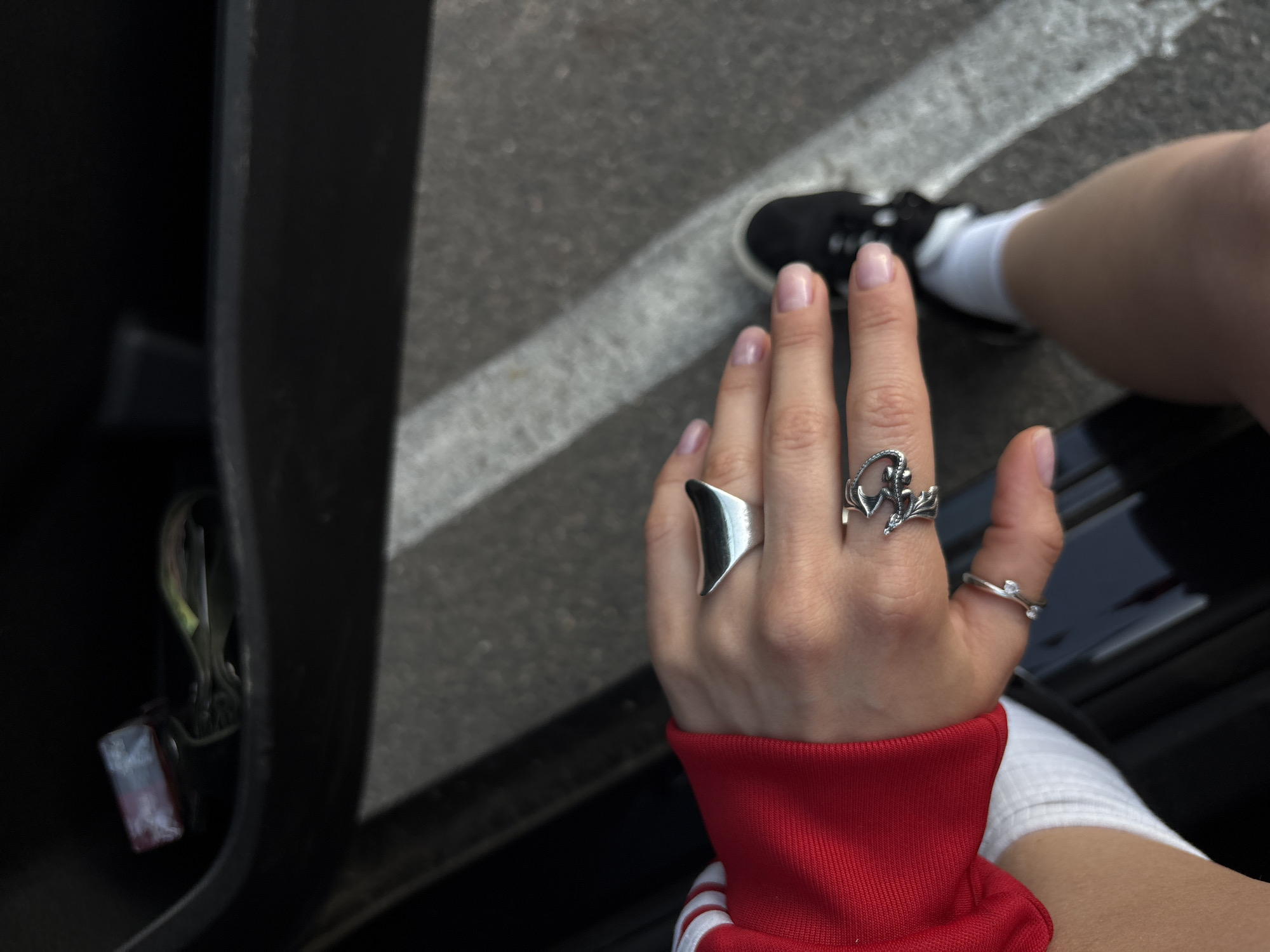 A person wearing a red Adidas track jacket and white socks sits with their legs visible, showcasing a hand adorned with three rings: a wide silver ring, a silver ring with a floral design, and a delicate silver ring with small stones. The background includes an open car door, a glimpse of a black shoe, and gray pavement with white lines. The location is indicated as Minsk, Belarus, although specific landmarks are not visible.