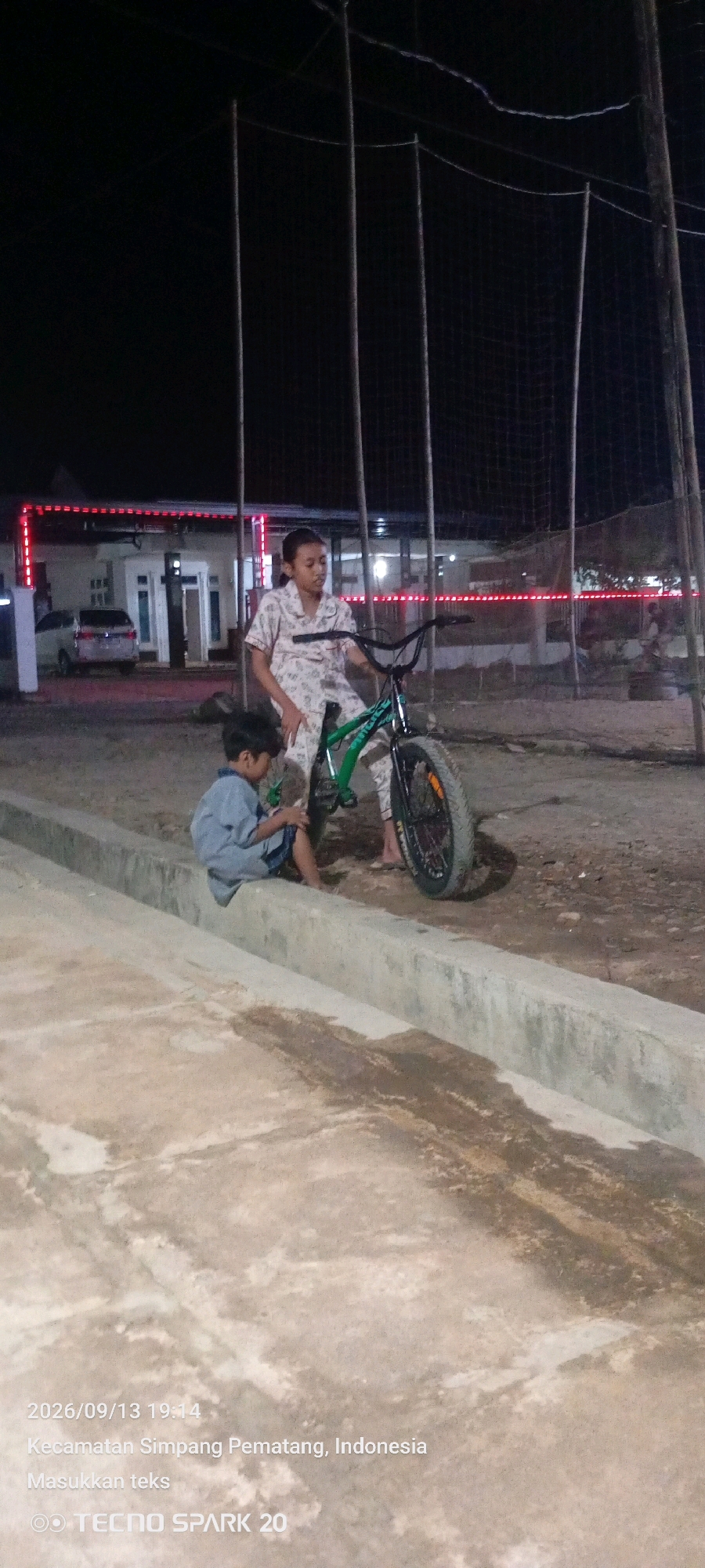 The image captures an outdoor scene at night in Kecamatan Simpang Pematang, Indonesia. Two children are present, with an older child, likely a girl, standing next to a green BMX bicycle. She is wearing a light-colored pajama set with a floral pattern. A younger child, dressed in a blue shirt and shorts, is seated on a concrete barrier, facing away from the camera and appearing to be interacting with something in his lap. Behind them, a building with red lights running along its facade is visible, with a white car parked in front. The background is dominated by tall poles and netting, suggesting an outdoor sports facility or enclosed area. The ground appears to be dirt or gravel. The timestamp in the lower left corner indicates the photo was taken on September 13, 2026, at 19:14.