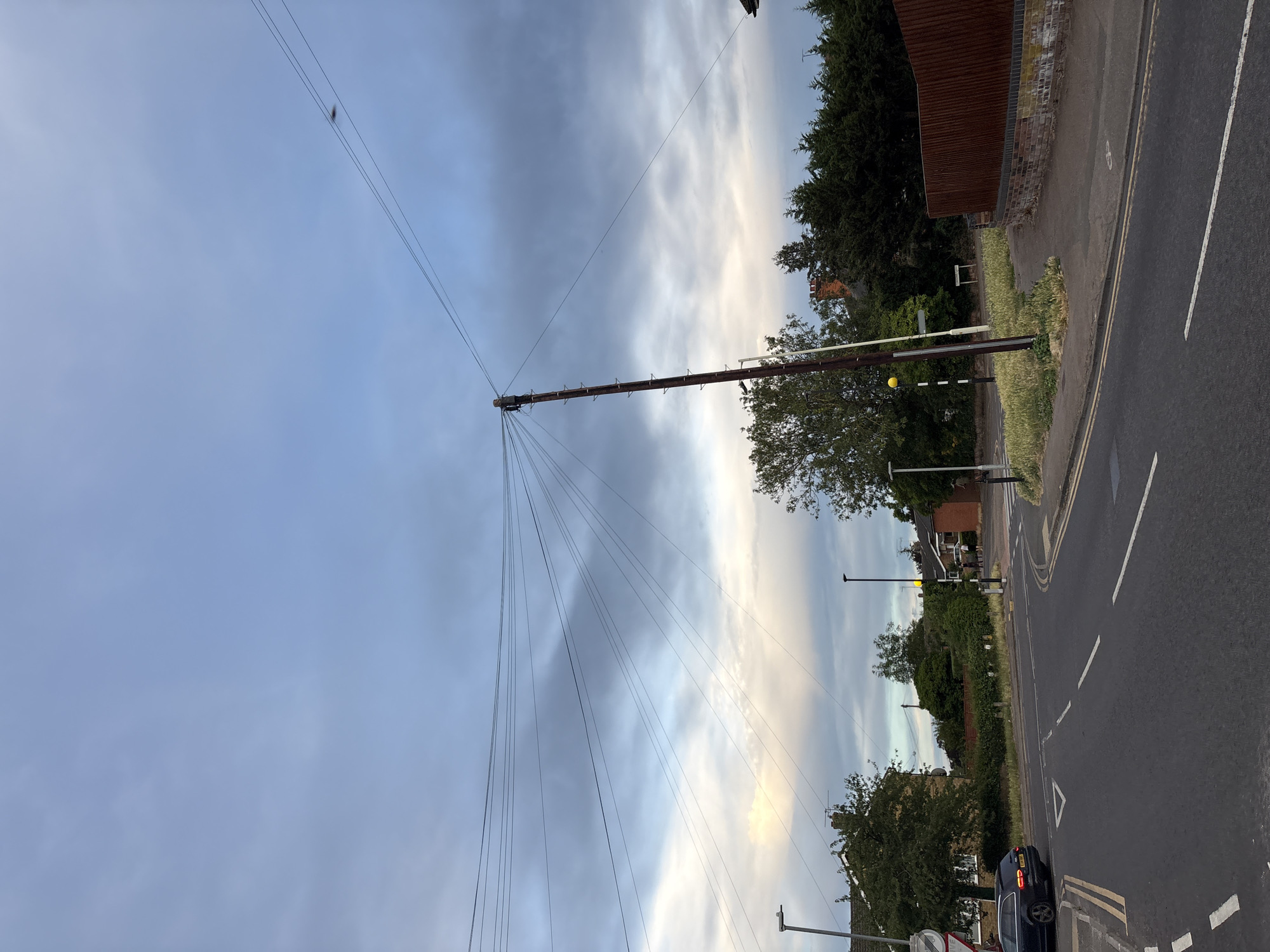 This image depicts a street scene in Chelmsford, United Kingdom, under a cloudy sky. The foreground is dominated by a road junction with clear lane markings. On the left side of the road, a dark colored car is visible. A large wooden utility pole with numerous wires extending from it stands prominently in the center of the image. In the background, houses, trees, and streetlights are present. The overall atmosphere suggests a residential area during daytime, possibly late afternoon or early evening, given the cloud cover and diffused light.