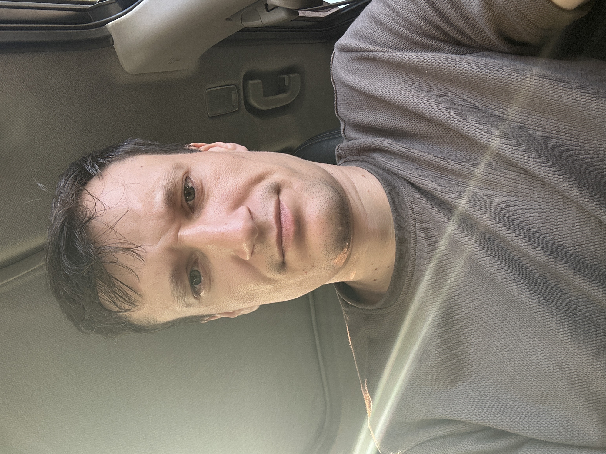 The image is a close-up selfie of a man inside a vehicle. He has short, dark brown hair and light-colored eyes. He is wearing a brown, textured t-shirt. Sunlight streams in from the left side of the frame, creating bright lines across his face and shirt. The interior of the vehicle is visible in the background, including a handle on the ceiling and the edge of a seat. There is no information in the image to suggest the location is Kamianets-Podilskyi, Ukraine.
