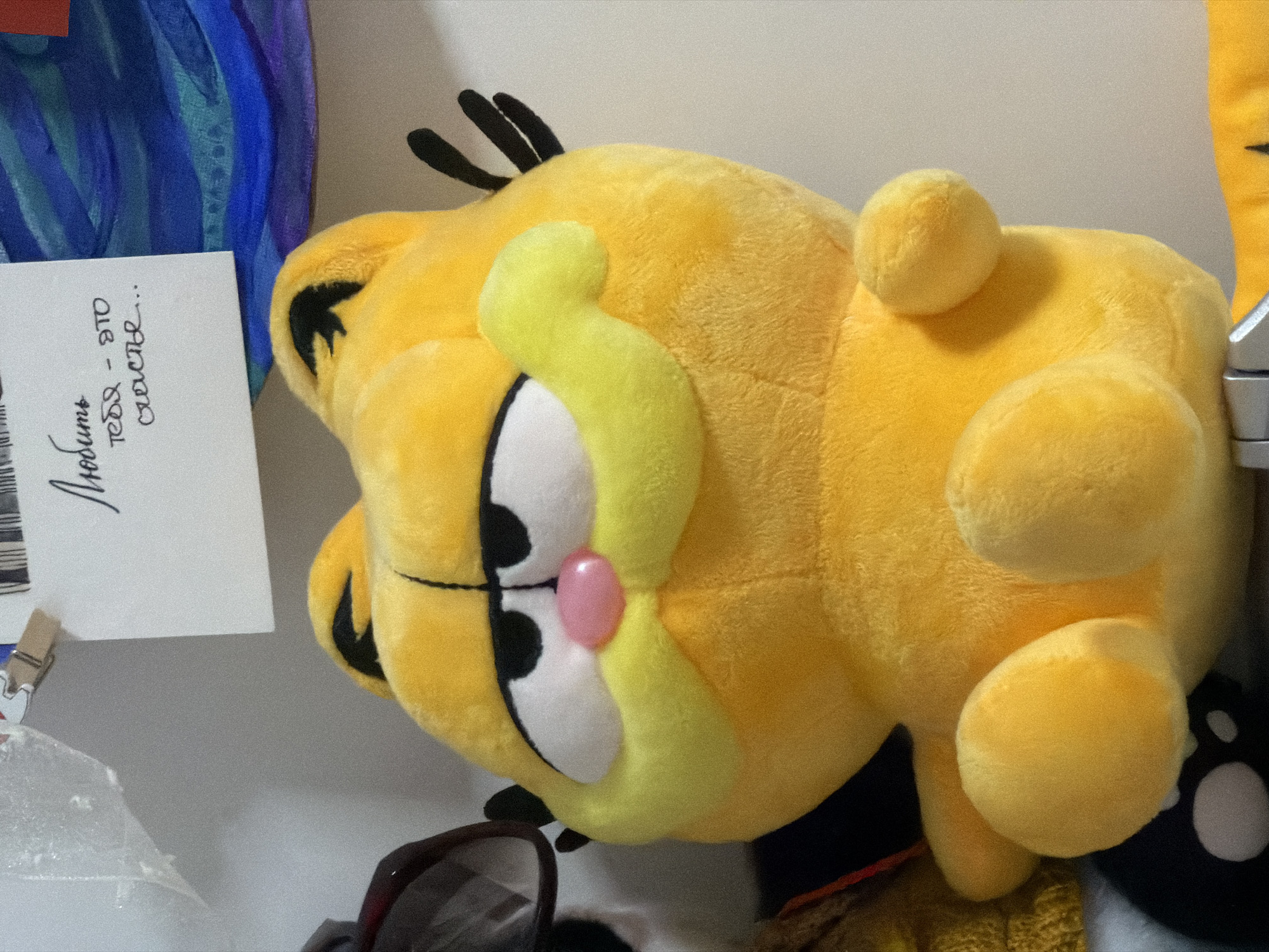 The image shows a Garfield plush toy lying on its side. A card with writing in Russian is clipped nearby. The text on the card is "Любить тебя - это счастье..." which translates to "Loving you - it's happiness...". The background is a neutral wall. The lighting suggests an indoor setting. A portion of what seems to be eyeglasses is visible. Without additional information, it's impossible to verify or deny that this photo was taken in Moscow, Russia.