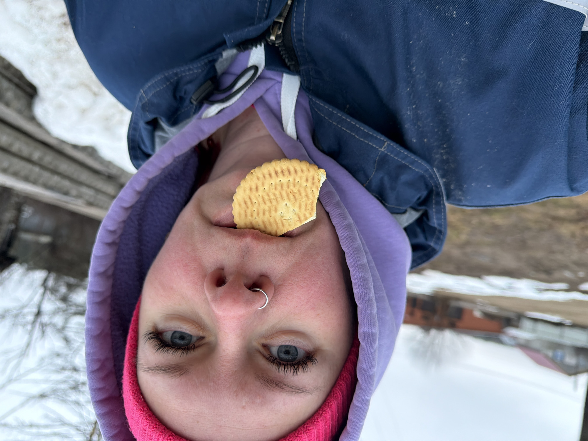 A young person with light skin and blue eyes is holding a cracker in their mouth, looking directly at the camera. They have a silver nose ring and are wearing a pink beanie under a purple hooded sweatshirt, which is layered under a blue jacket with a zipper. The background includes snow, trees, and what appears to be a building, suggesting an outdoor environment in a residential area. The location is Chaikovsky, Russia.
