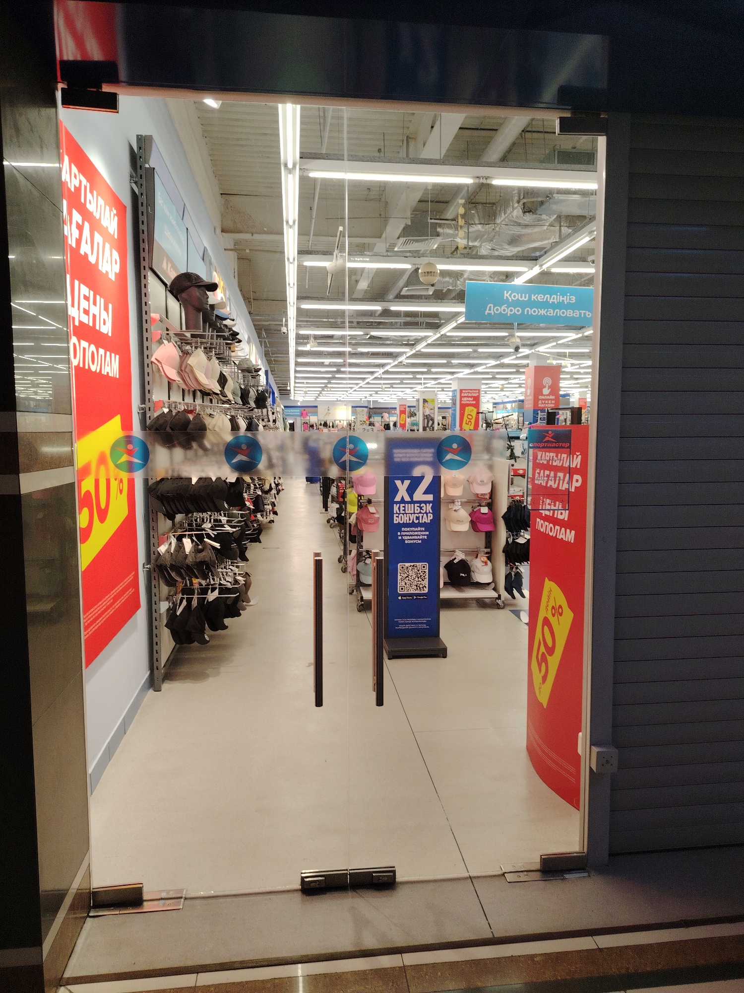 This is a medium-shot, eye-level, exterior view of a retail store entrance, likely in Astana, Kazakhstan, as indicated by the Cyrillic text on the signage. The time of day appears to be evening or dusk, judging by the interior lighting being significantly brighter than any ambient exterior light, and the closed security shutters on the right side of the frame.
The scene is dominated by a glass-fronted entrance to a store, with a large red banner on the left side featuring white Cyrillic text that reads "АРТЫЛАЙ АҒАЛАР ЦЕНЫ ОПОЛАМ" and a yellow graphic with "50%". This suggests a sale or discount event. Inside the store, racks of hats and caps are visible, displayed on metal shelving. Another red banner, similar to the one outside, is visible further inside the store, also advertising "50%". A blue sign with white Cyrillic text, "Қош келдіңіз Добро пожаловать," meaning "Welcome," is also visible inside, reinforcing the retail setting.
On the left side of the entrance, there is a display with a QR code and text indicating "X2 КЕШБЭК БОНУСТАР ПОКУПАЙТЕ В ПРИЛОЖЕНИИ И УДВАИВАЙТЕ БОНУСЫ," which translates to "X2 Cashback Bonuses. Buy in the app and double your bonuses." This highlights a promotional activity. The store appears to be a sports apparel and accessories retailer, possibly "Спортмастер" (Sportmaster), as suggested by a visible logo. The floor is a light grey, and the ceiling has a grid of fluorescent lights. The overall impression is that of a modern, well-lit retail environment. No people are clearly visible within the store itself, but the mannequins displaying hats suggest the presence of merchandise. The glass doors are a focal point, reflecting some of the interior lights and signs.