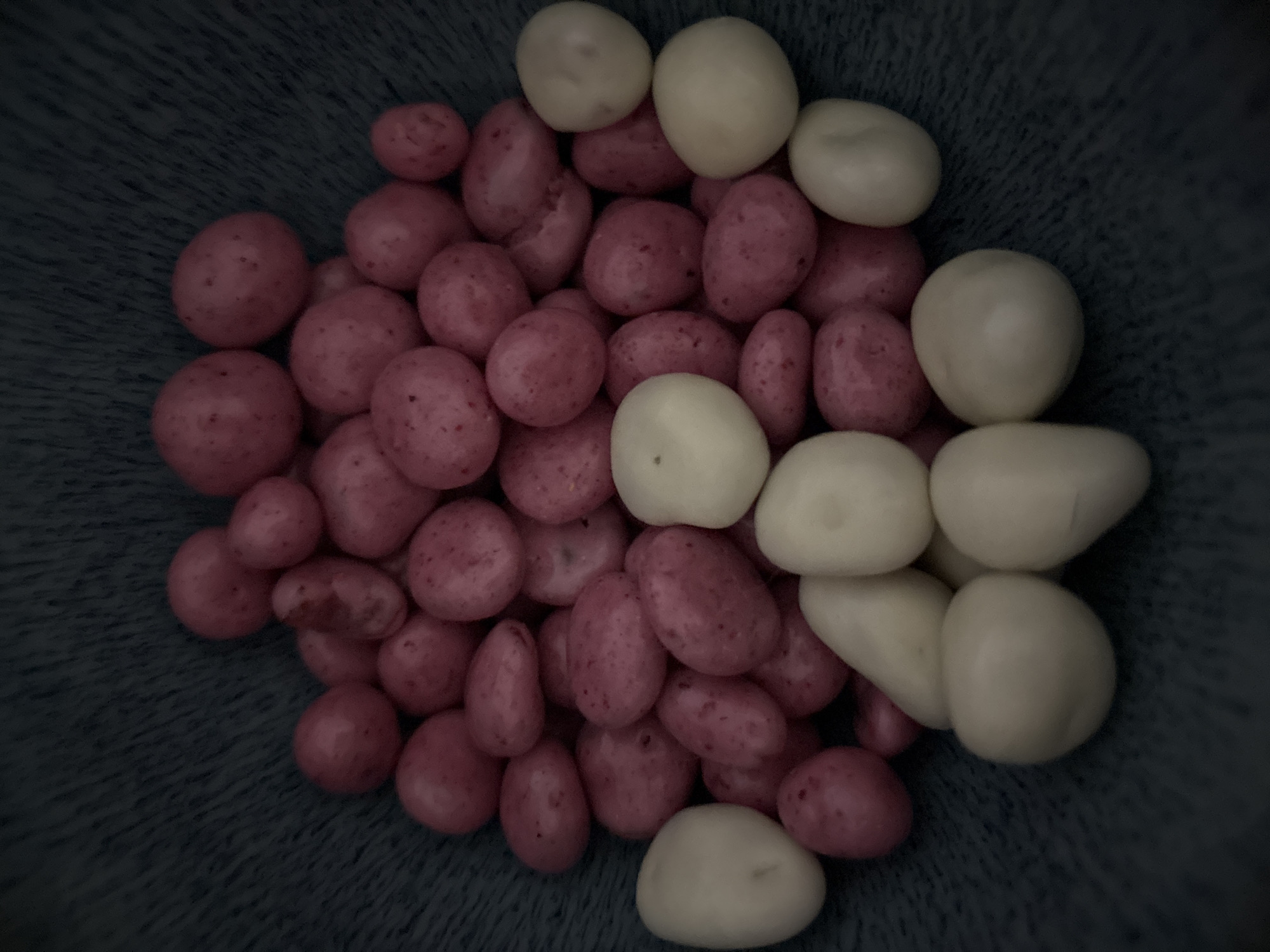 A close-up, overhead shot shows a pile of small, pink, oblong candies mixed with a few larger, white, oval candies in a dark blue bowl. The candies are clustered together, filling most of the frame. The background is the textured surface of the bowl, which appears to be made of ceramic or a similar material. The lighting is dim, creating shadows and highlighting the smooth surfaces of the candies.