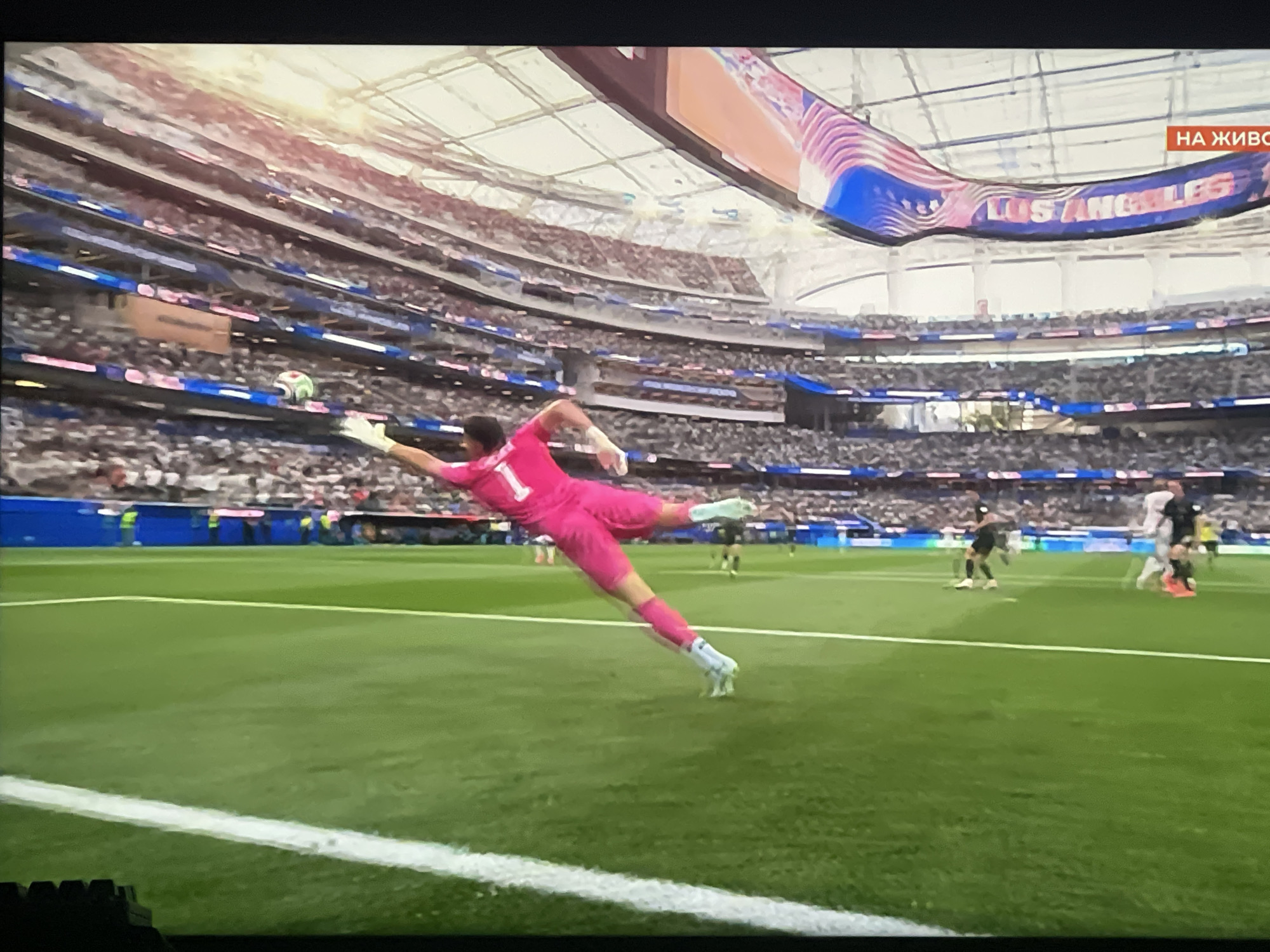 A soccer goalkeeper in a pink uniform dives to block a ball during a match inside a brightly lit stadium. The goalkeeper's body is stretched out, with one leg extended and the other bent at the knee. The goalkeeper's hands are reaching towards the ball, which is a green and white soccer ball visible in the air. The field is a vibrant green, with white lines marking the boundaries. In the background, a large crowd of spectators fills the stadium seats, and a large screen displays graphics and text, including "LOS ANGELES" in large letters. The stadium has a modern, domed roof. There is a sign in Russian that says "ON AIR". The scene takes place in a professional sports stadium, likely during a professional soccer match. The location is not Ruse, Bulgaria.