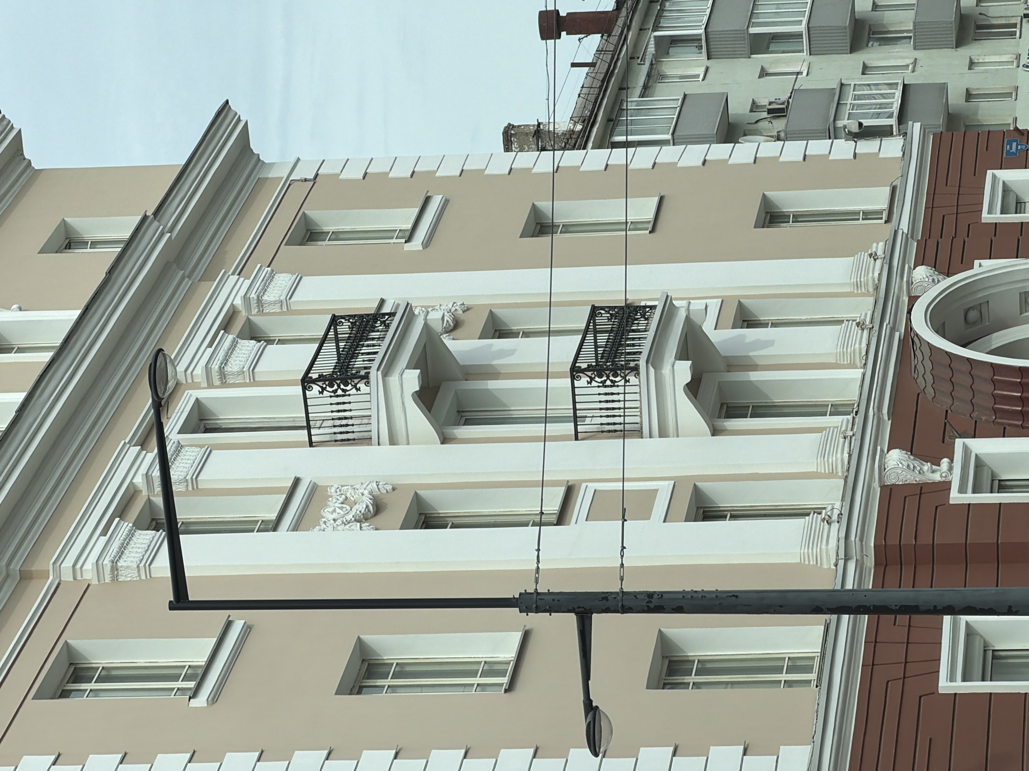 A low-angle shot captures the exterior of a multi-story building in Perm, Russia. The building has a predominantly beige facade with white trim outlining the windows and architectural details. Some windows feature black fire escapes. A utility pole and streetlight are visible in the foreground. Adjacent to the building is another structure with a brown facade and a rounded architectural element. In the background, a taller building with visible air conditioning units on its exterior can be seen. The sky is overcast.