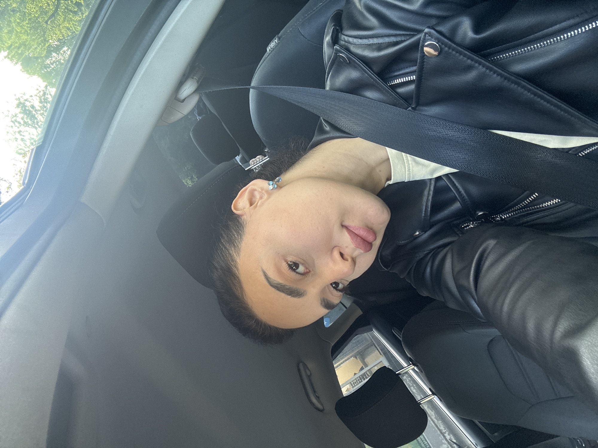 A woman wearing a black leather jacket and secured with a seatbelt is in a car. The location is likely in the city of Tbilisi, Georgia, based on the outside view seen through the car window. The photo is taken at an unusual angle. The woman is looking toward the camera.